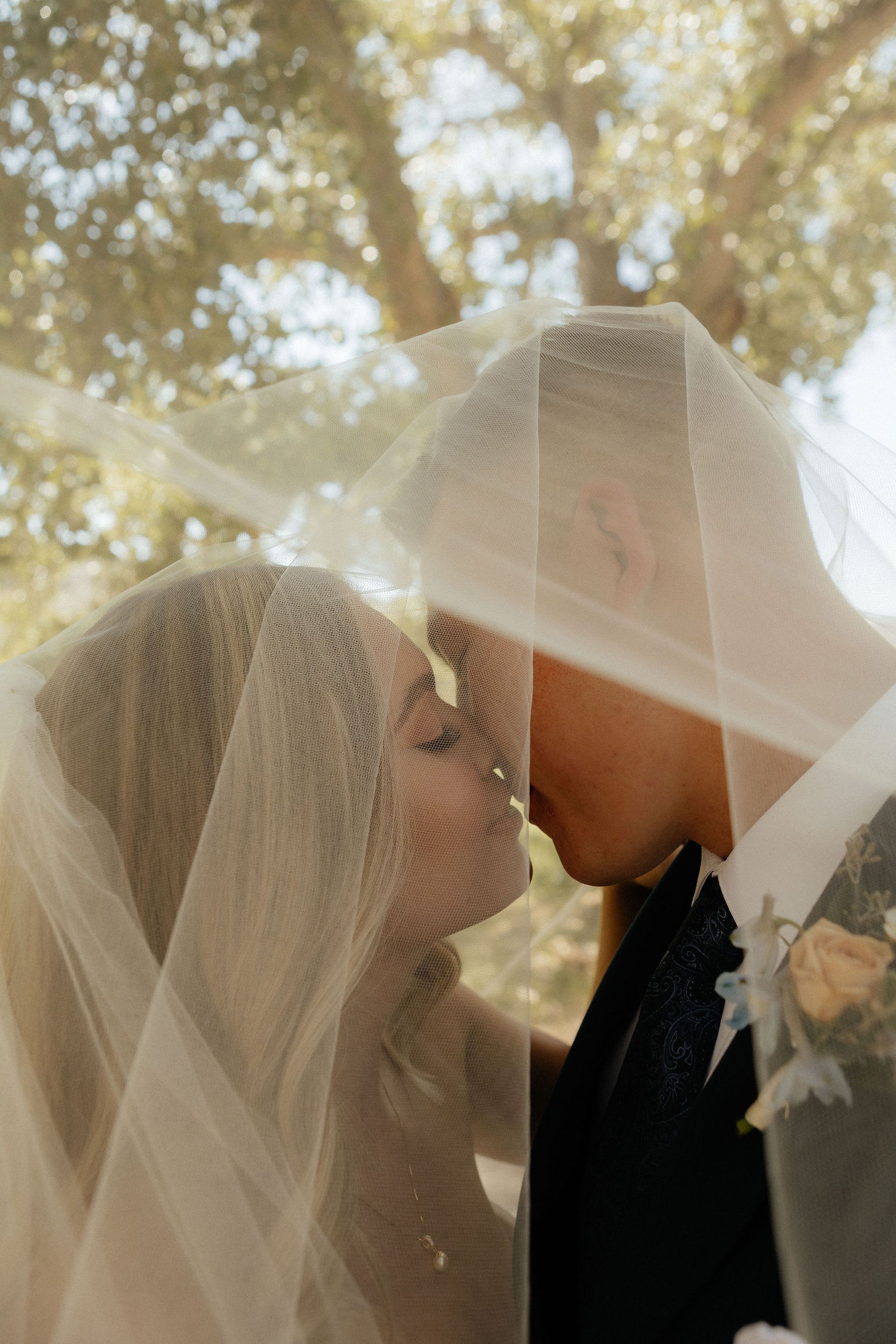 Bride and groom kissing under a sheer veil outdoors with trees in the background