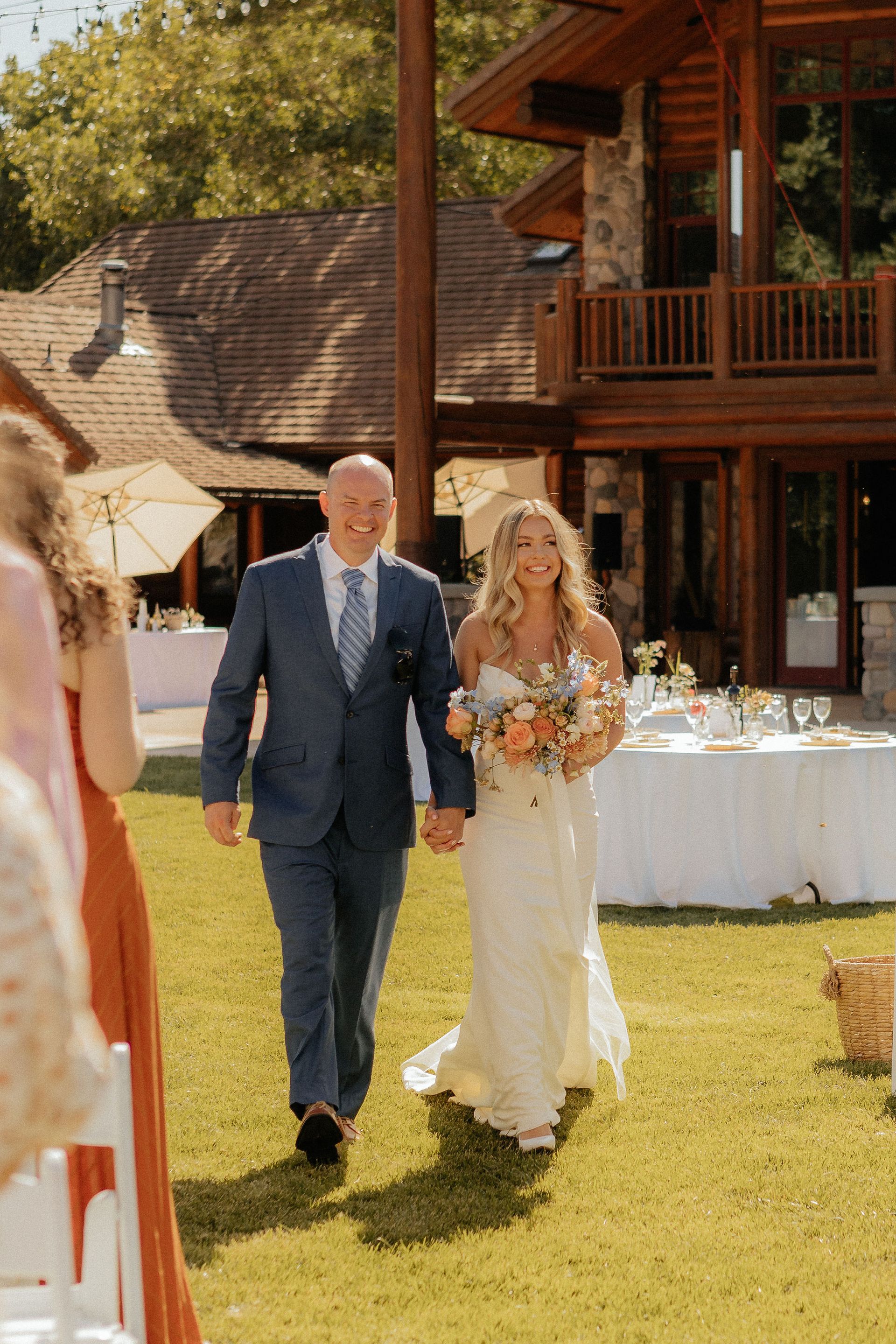 Bride and older man walking on grass at an outdoor wedding reception, with a rustic wooden building behind them.