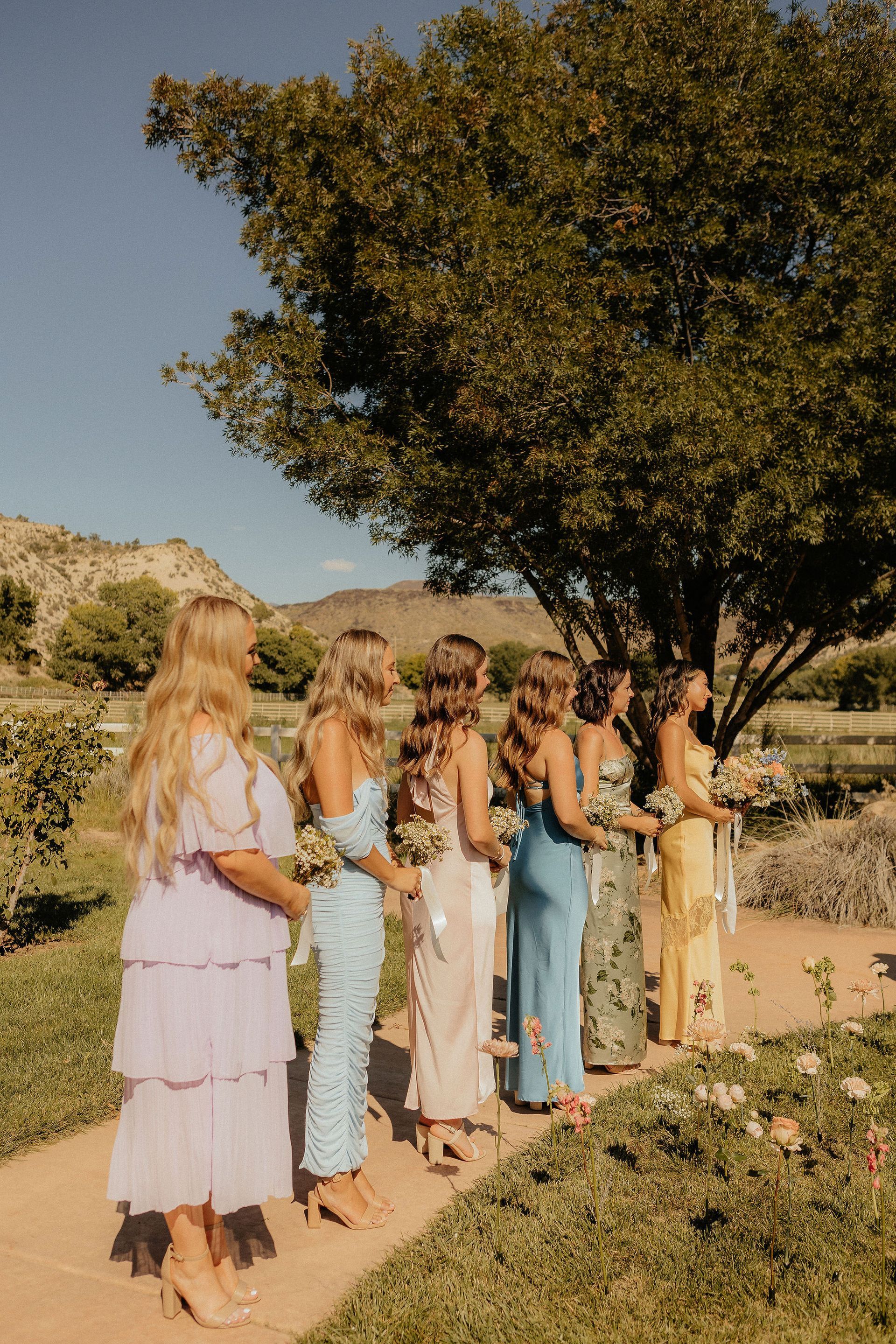 Women in pastel dresses standing outdoors in a garden under a large tree, holding bouquets.