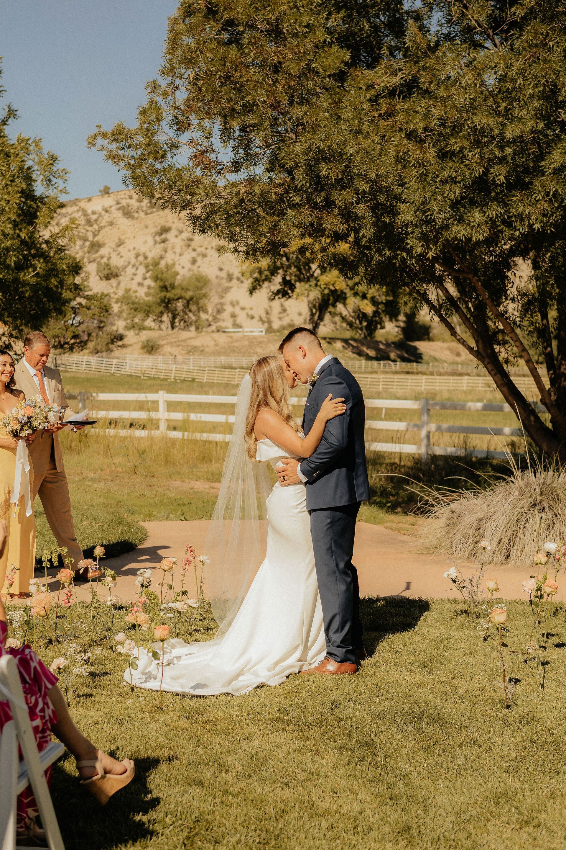 Couple embracing during outdoor wedding ceremony under a tree, with guests nearby and a rural landscape behind them.