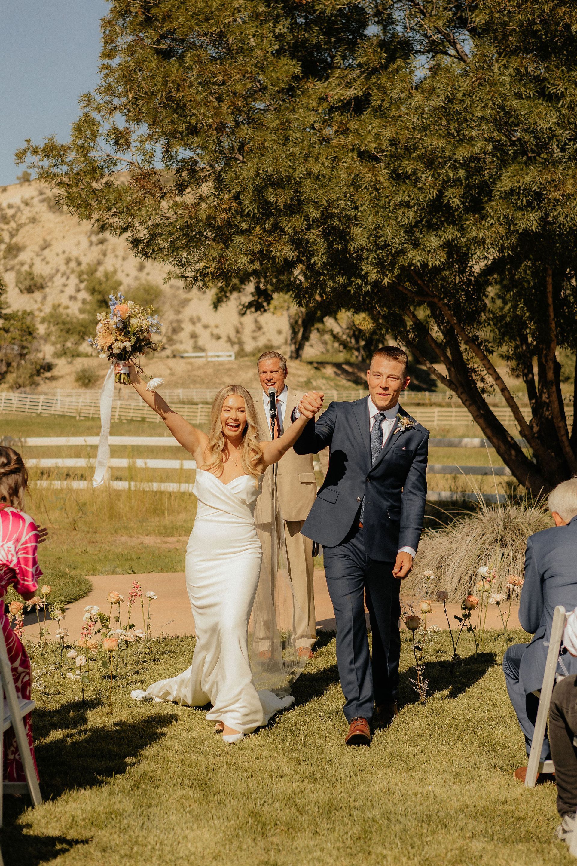 Bride and groom walking down an outdoor aisle, smiling and raising their hands in celebration.