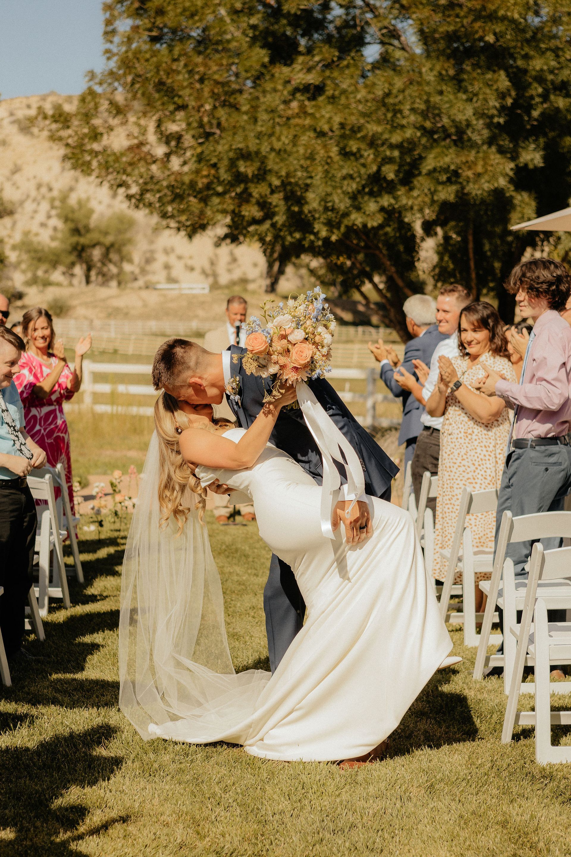 Wedding couple kissing outdoors under a tree as guests clap nearby
