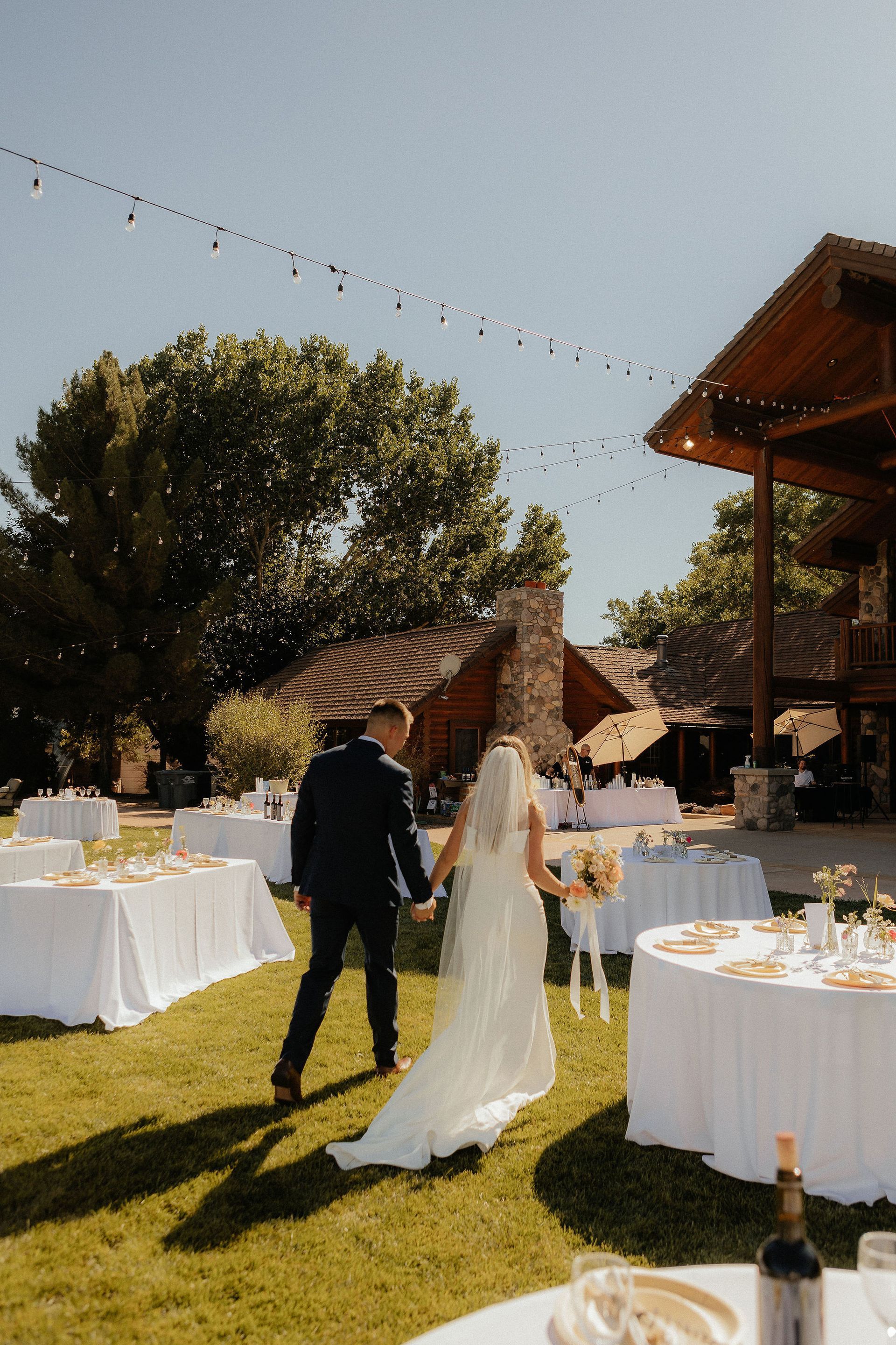 Couple walking hand in hand through an outdoor wedding reception with white-clothed tables and string lights.