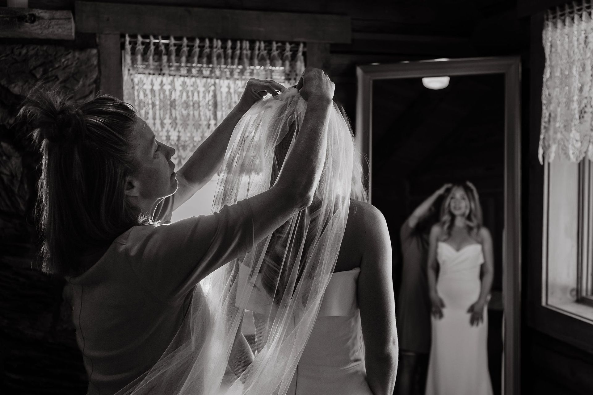 Bridesmaid adjusts a bride’s veil as the bride stands in a doorway, looking on.
