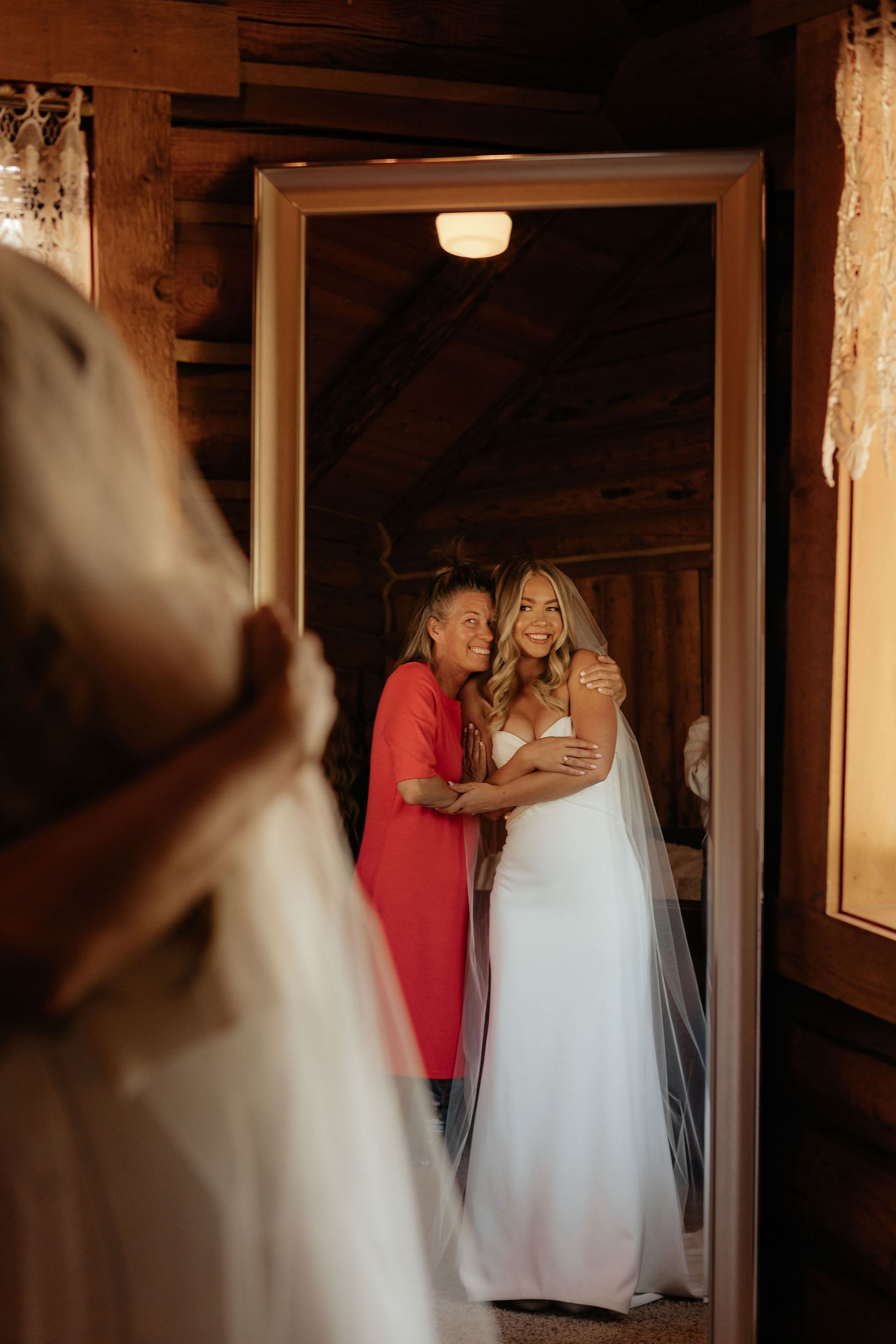 Two women in formal dresses embrace in a wooden doorway, one in white and one in red, with a veil in the foreground