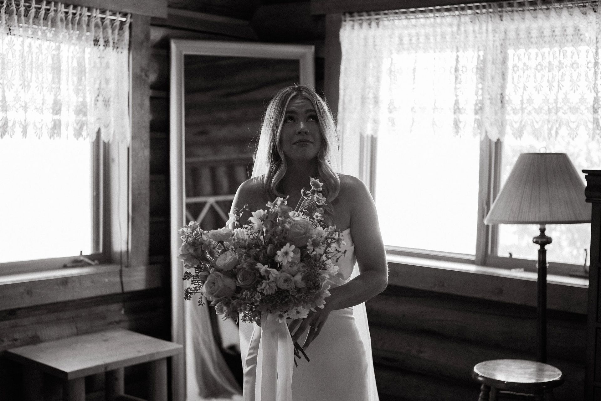Bride holding a bouquet in a rustic room by curtained windows and a floor lamp