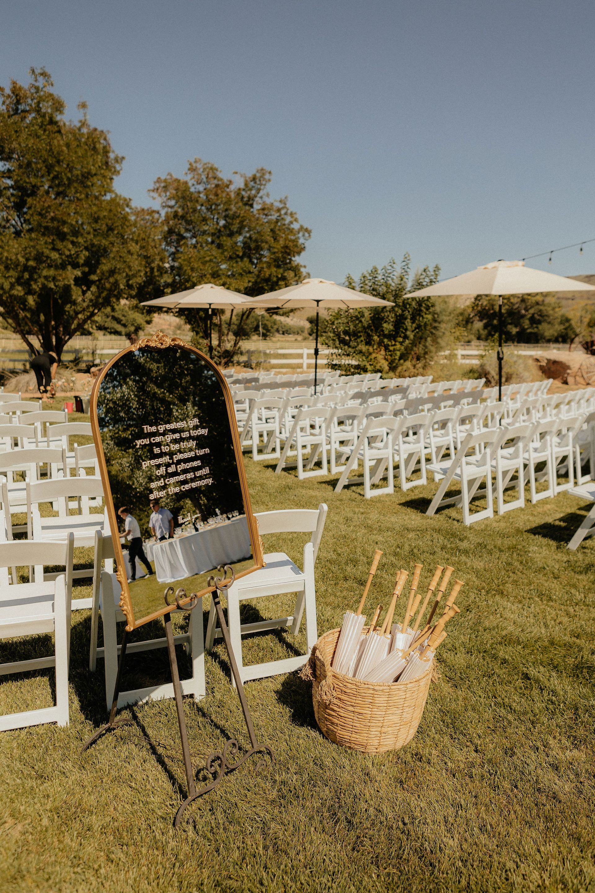 Outdoor wedding setup with white chairs, a chalkboard sign, and a wicker basket on grass under umbrellas