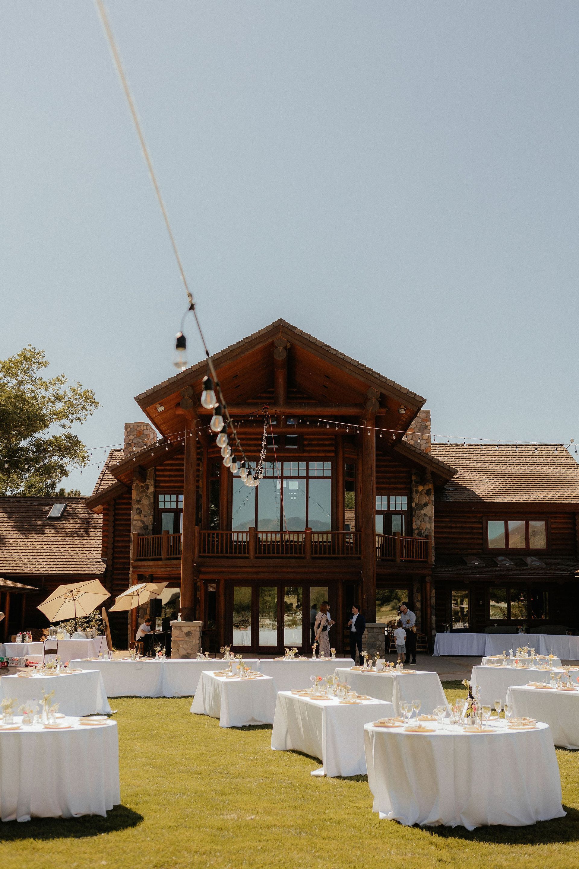 Outdoor wedding reception in front of a rustic wooden lodge with white tables on a grassy lawn