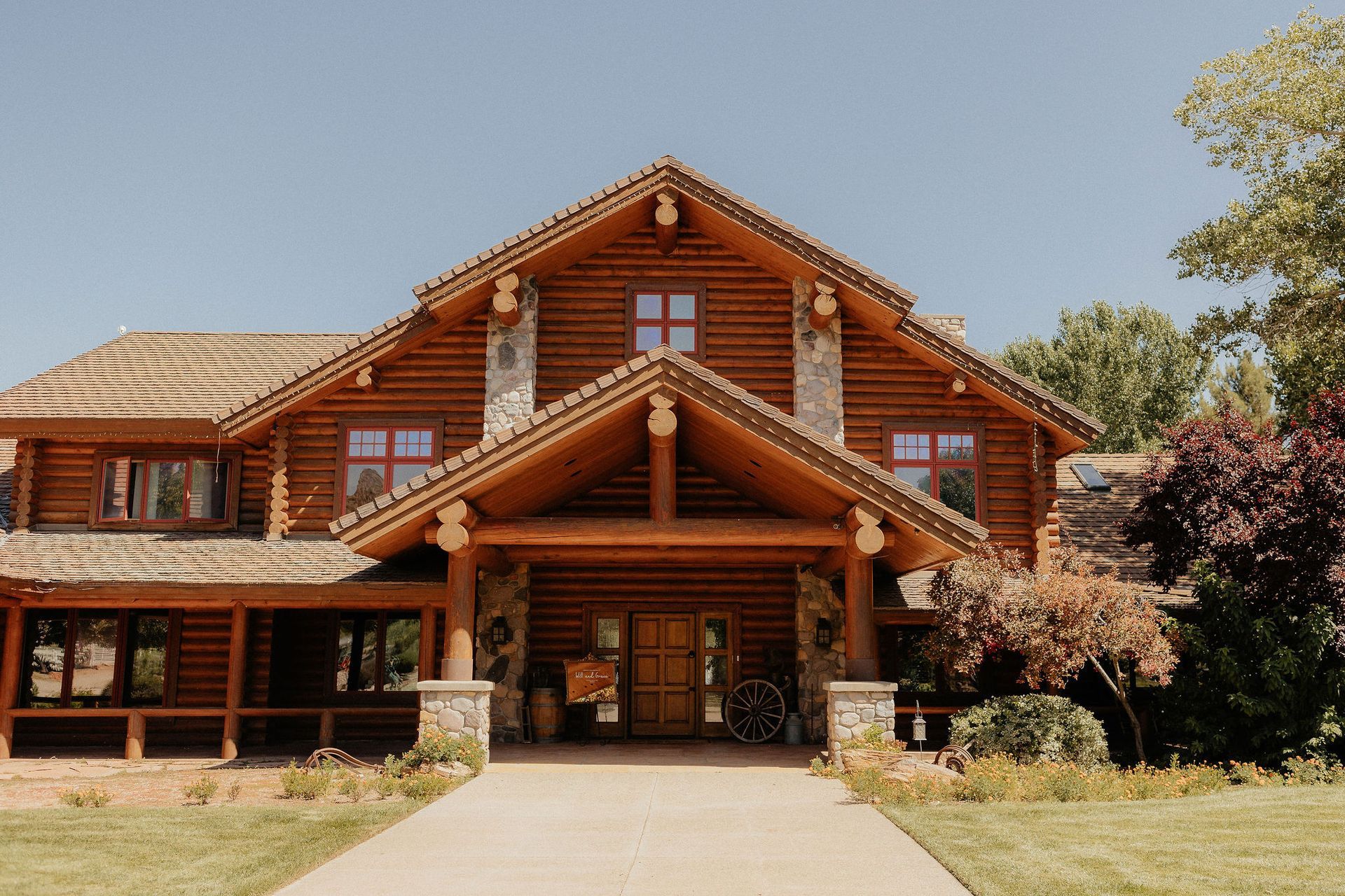 Large rustic log cabin with a covered porch and central stone entrance, surrounded by trees and lawn.