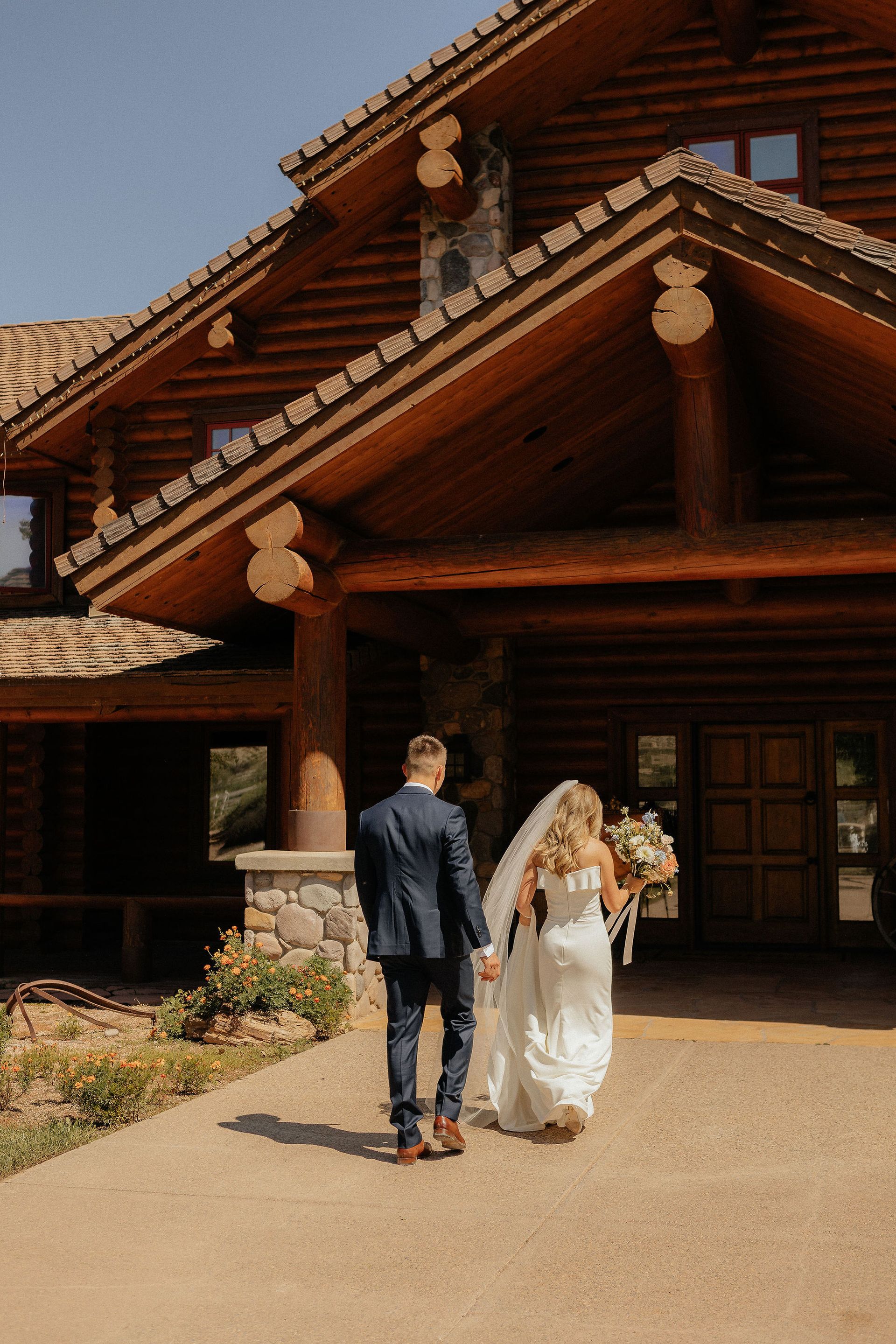 Couple walking toward a rustic log cabin entrance, holding flowers and dressed for a wedding.