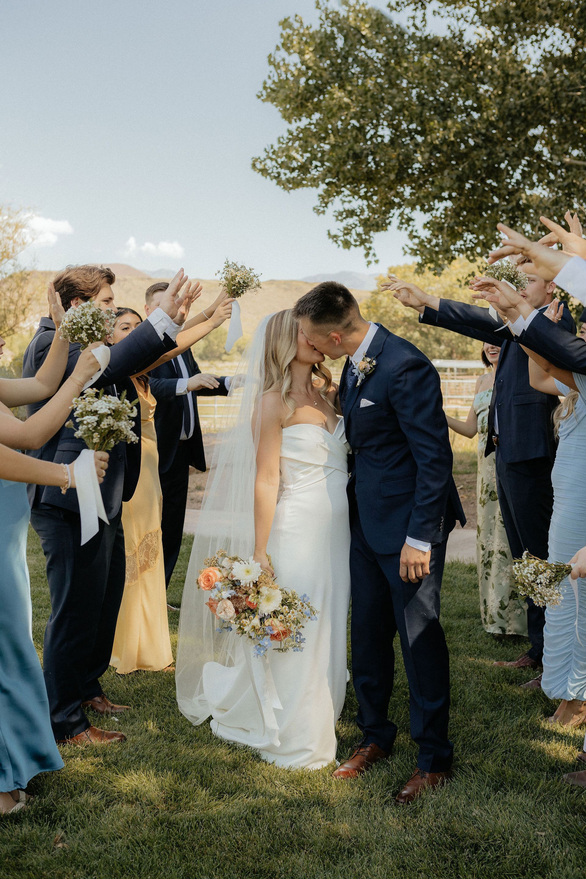 Wedding couple kissing under an arch of raised arms outdoors, bride in white gown, groom in navy suit