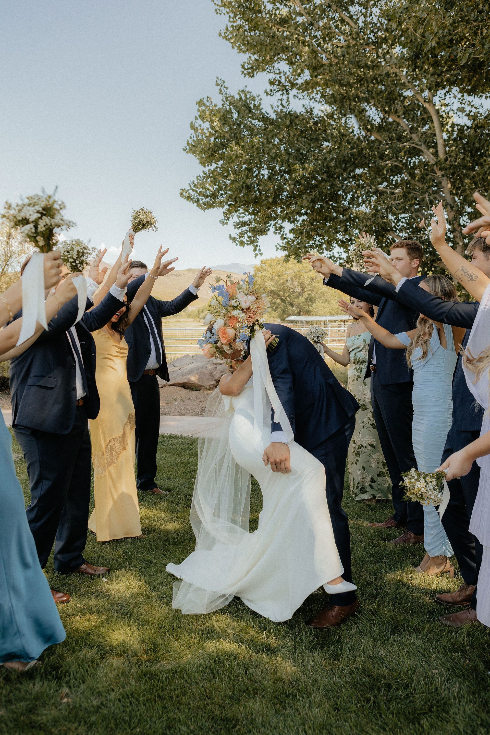 Wedding party cheering as the bride and groom kiss outdoors under a tree