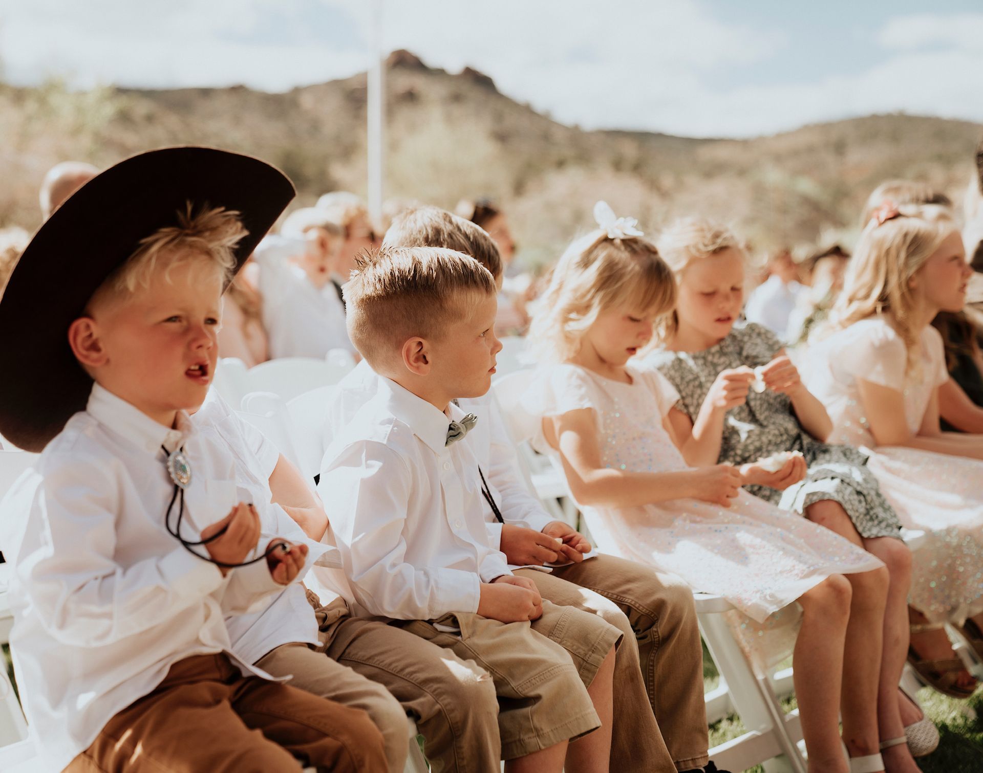 Children in white clothes and hats sitting outdoors on white chairs, holding snacks at a sunny event