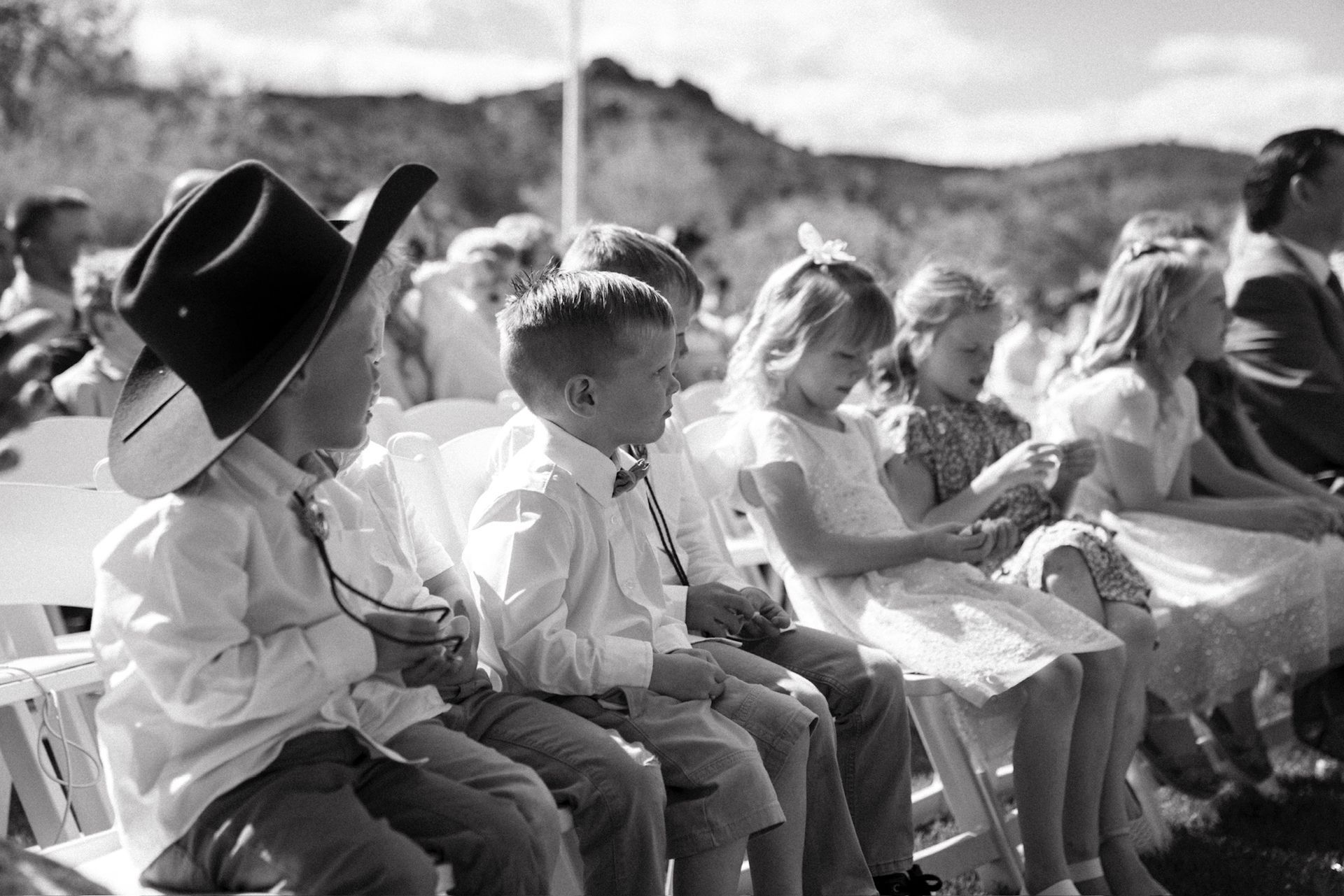 Children seated in white chairs at an outdoor event, one wearing a large cowboy hat