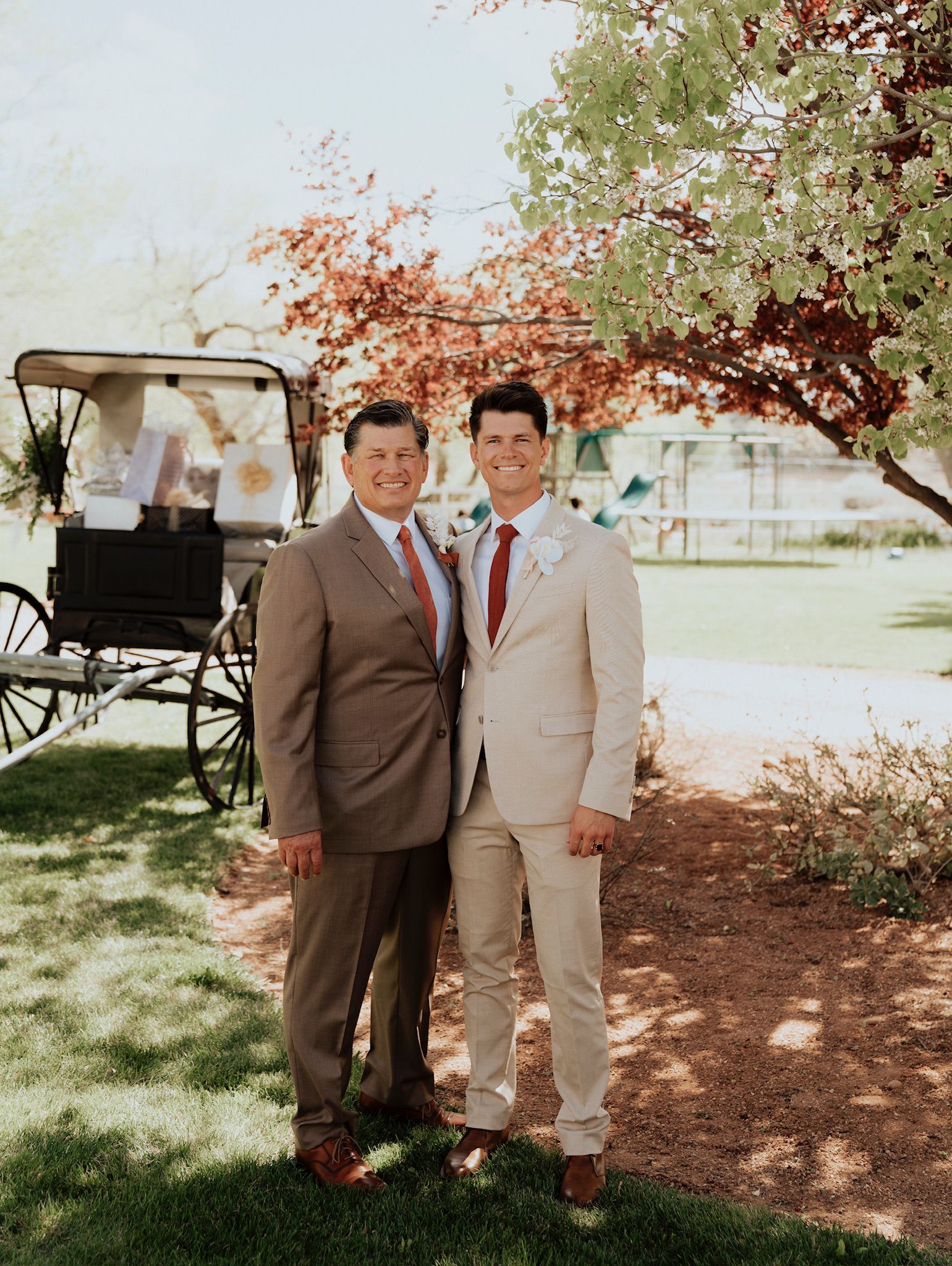 Two men in suits standing outdoors beside a horse-drawn carriage under a tree with autumn leaves
