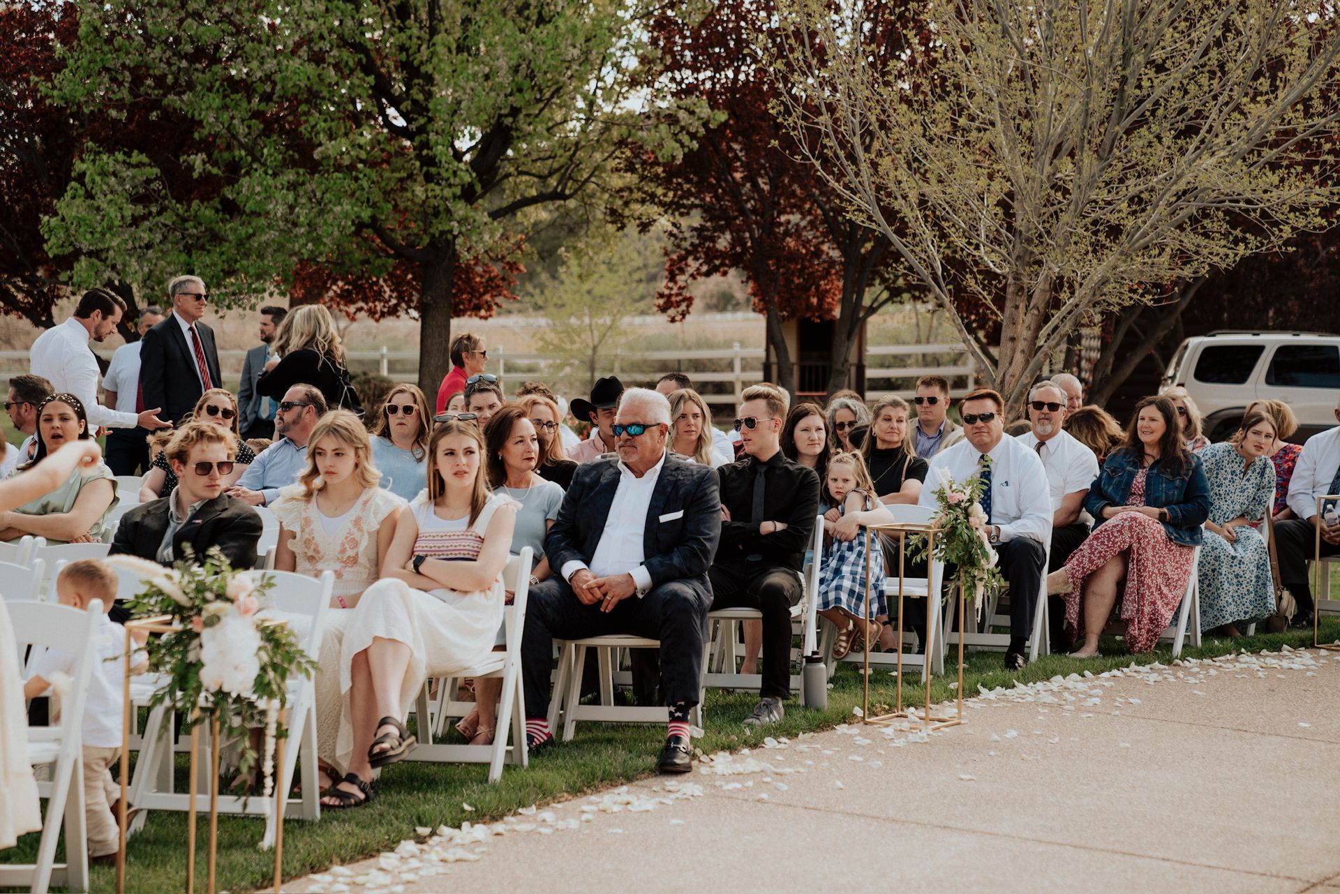 Outdoor wedding guests seated in white chairs under trees, with the bride and groom in the front row