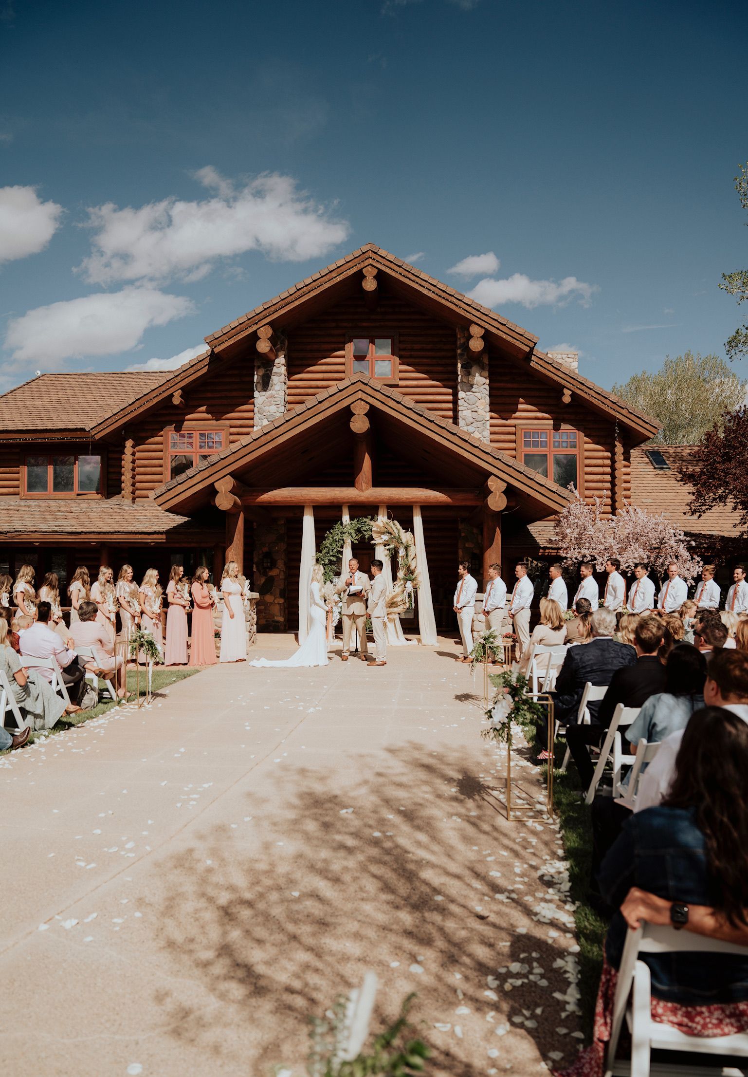 Wedding party posed outside a log cabin chapel, with guests lining the path under a bright blue sky.