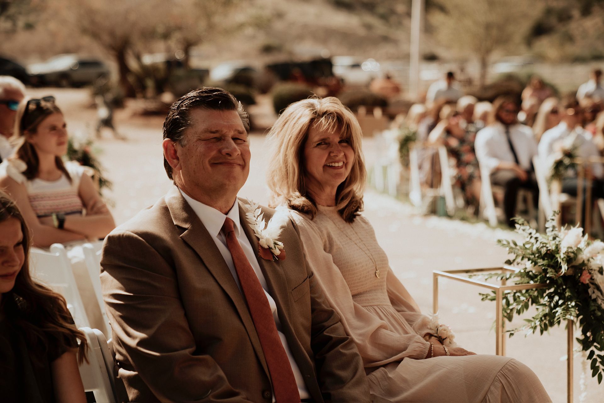 Seated guests at an outdoor ceremony, smiling in warm sunlight with floral décor nearby