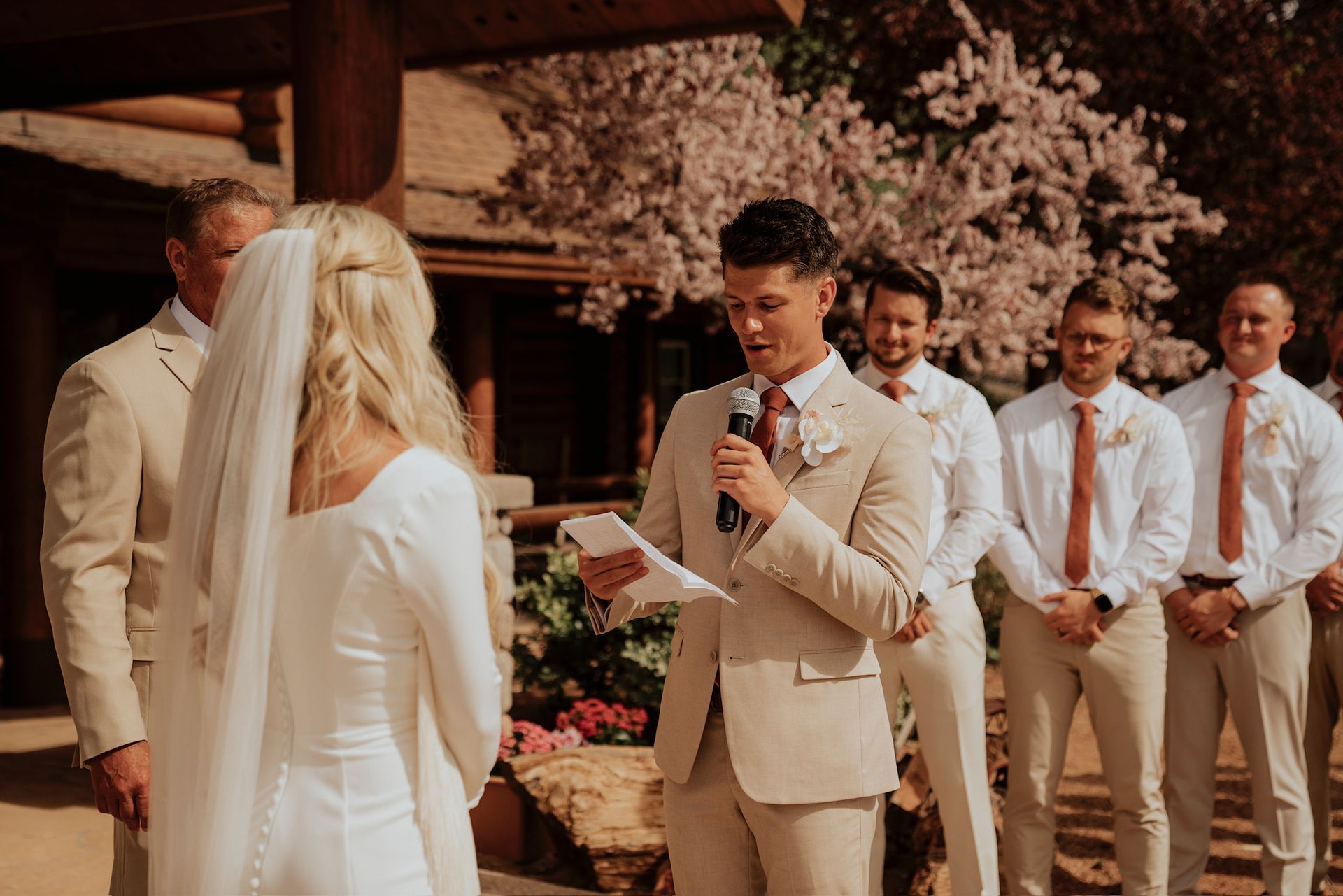 Wedding ceremony under a wooden pavilion, with a man speaking to the couple and groomsmen standing nearby