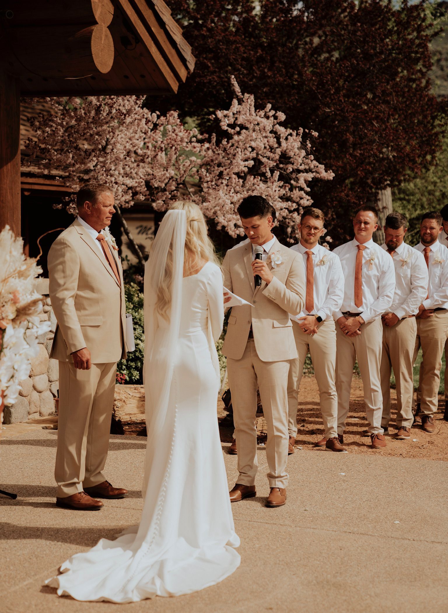 Wedding ceremony outdoors with bride and groom standing before attendants in cream suits