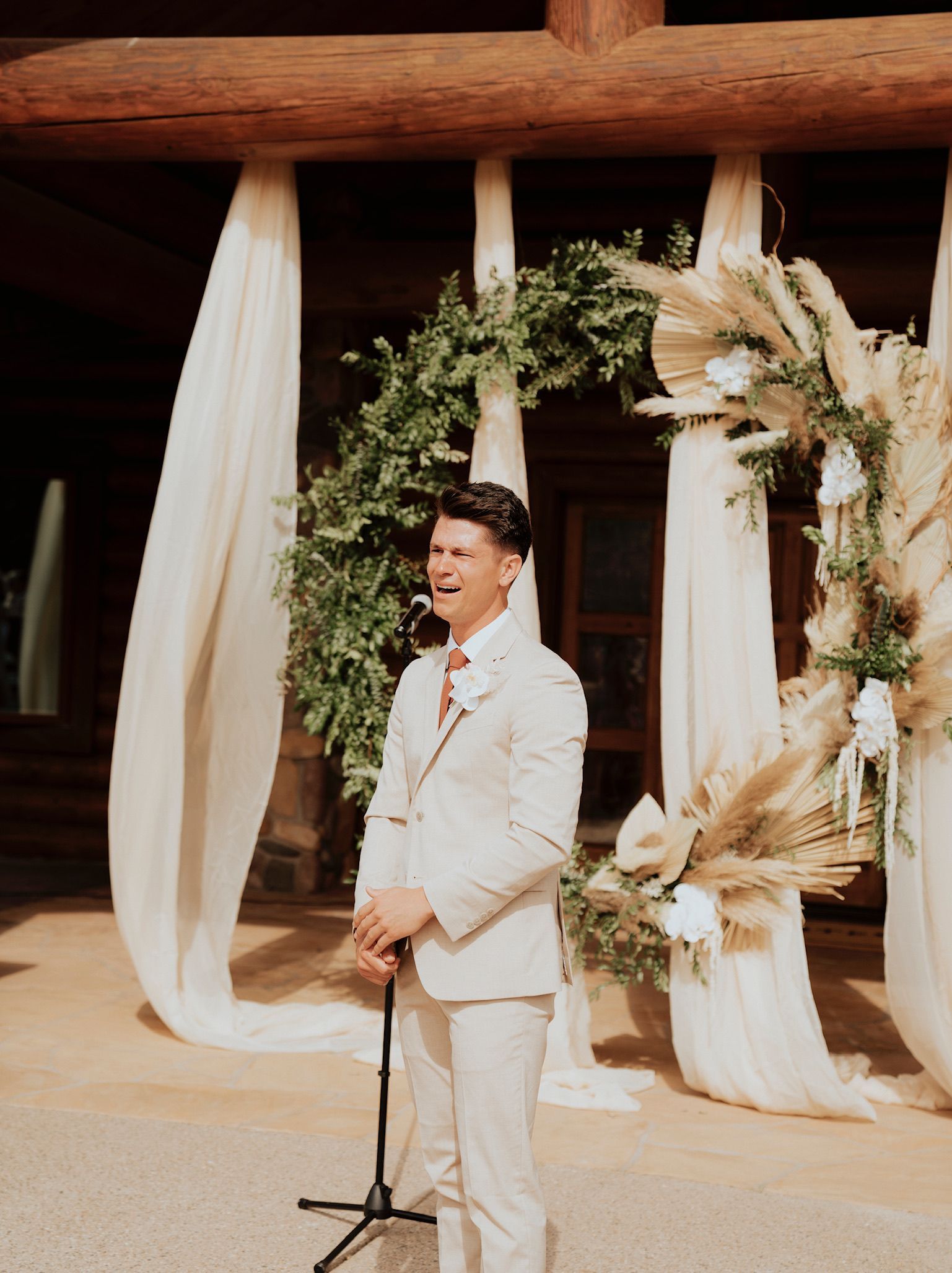 Man in a white suit speaking at a microphone beneath a decorated floral arch outdoors