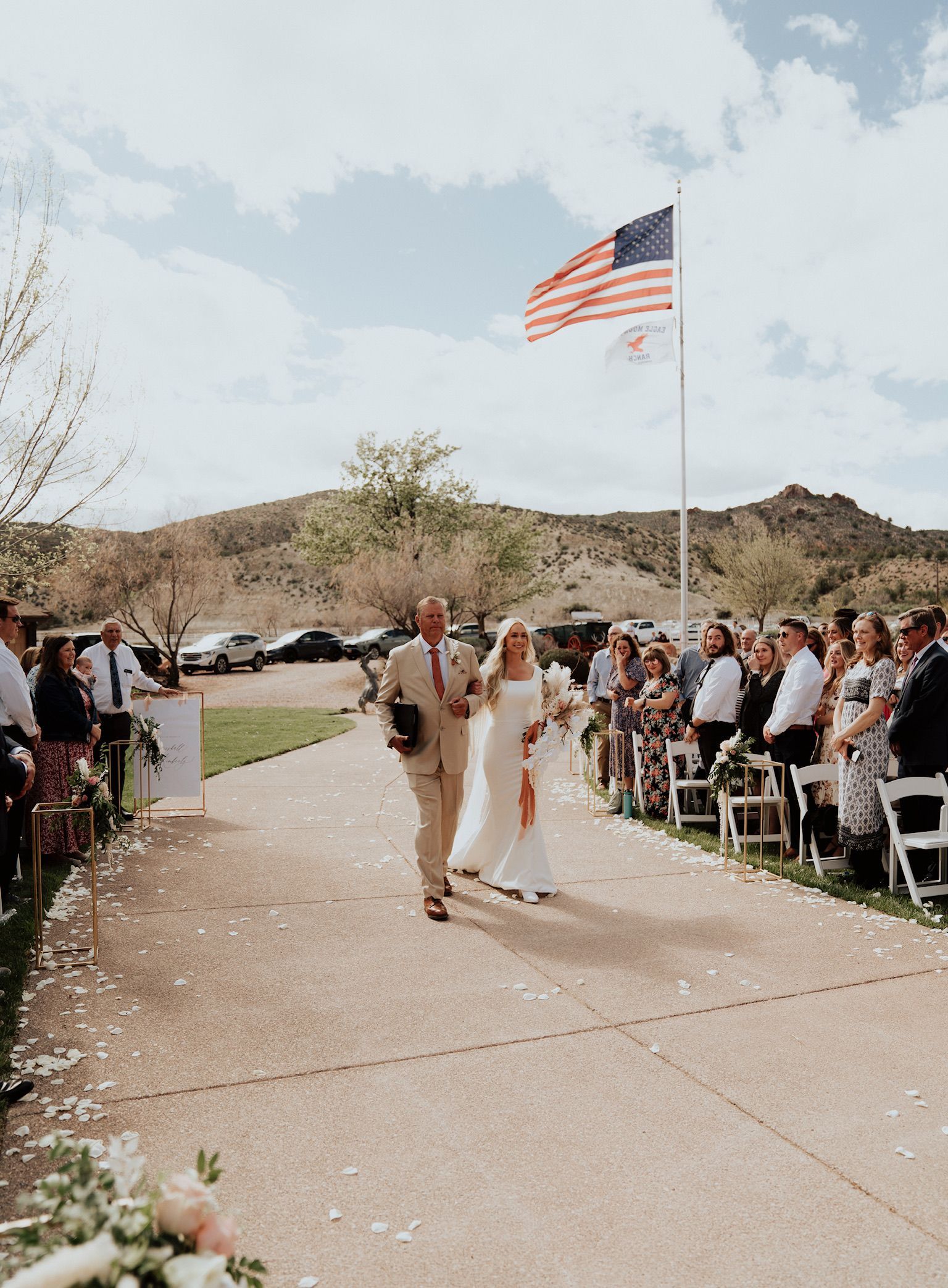 Outdoor wedding ceremony with couple walking down an aisle under a large American flag.