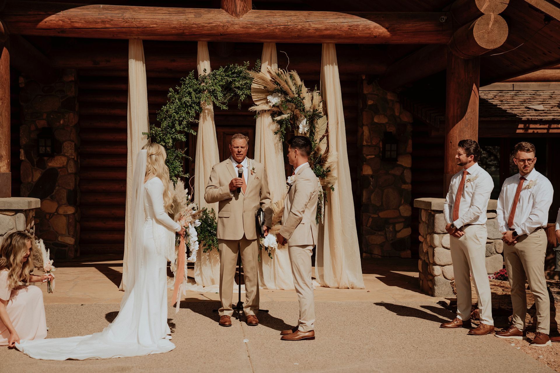 Wedding ceremony outside a rustic log cabin, with bride, groom, officiant, and attendants standing in the sun.