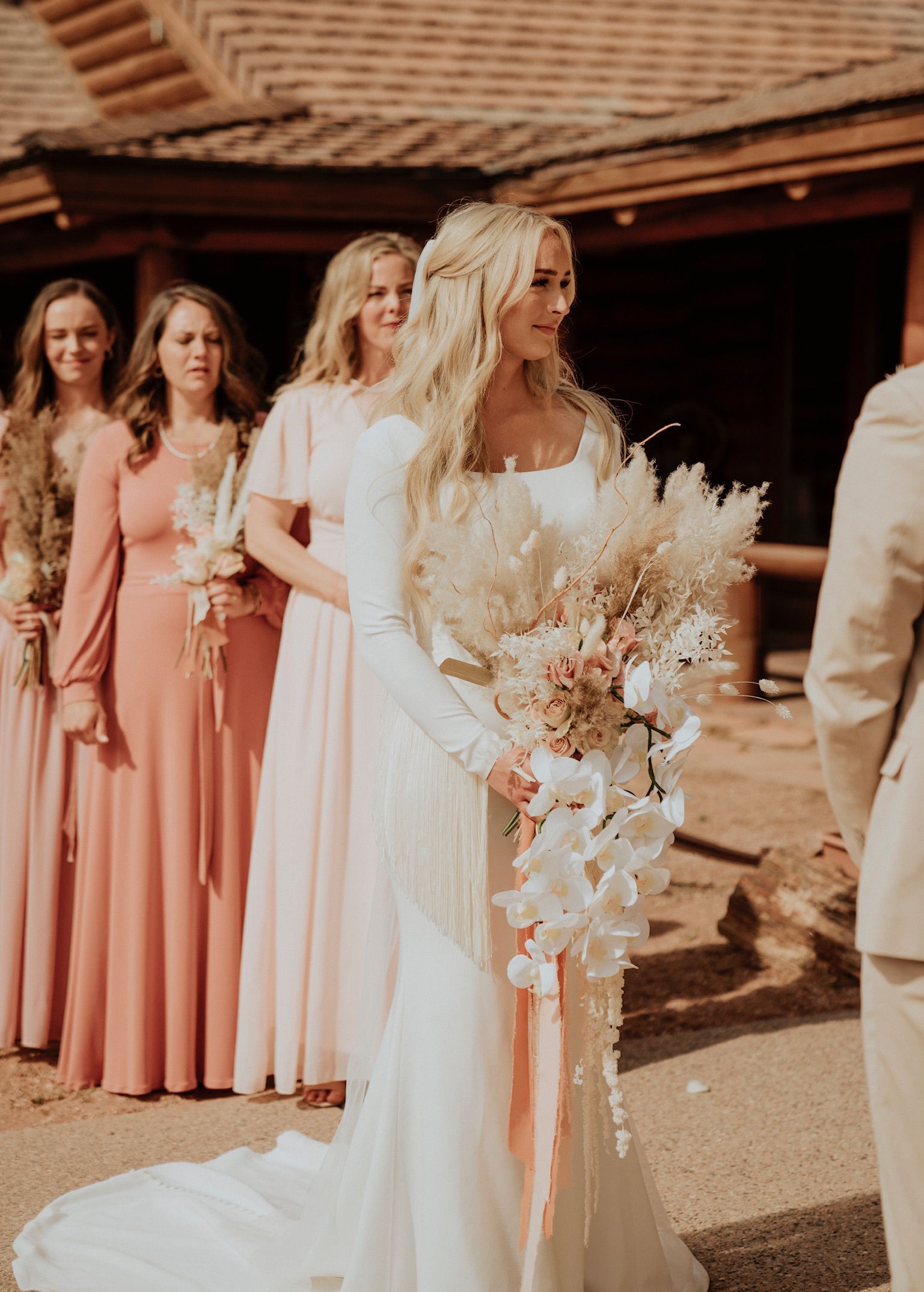 Bridal party in blush dresses and white gowns standing outdoors, holding a bouquet at a rustic venue