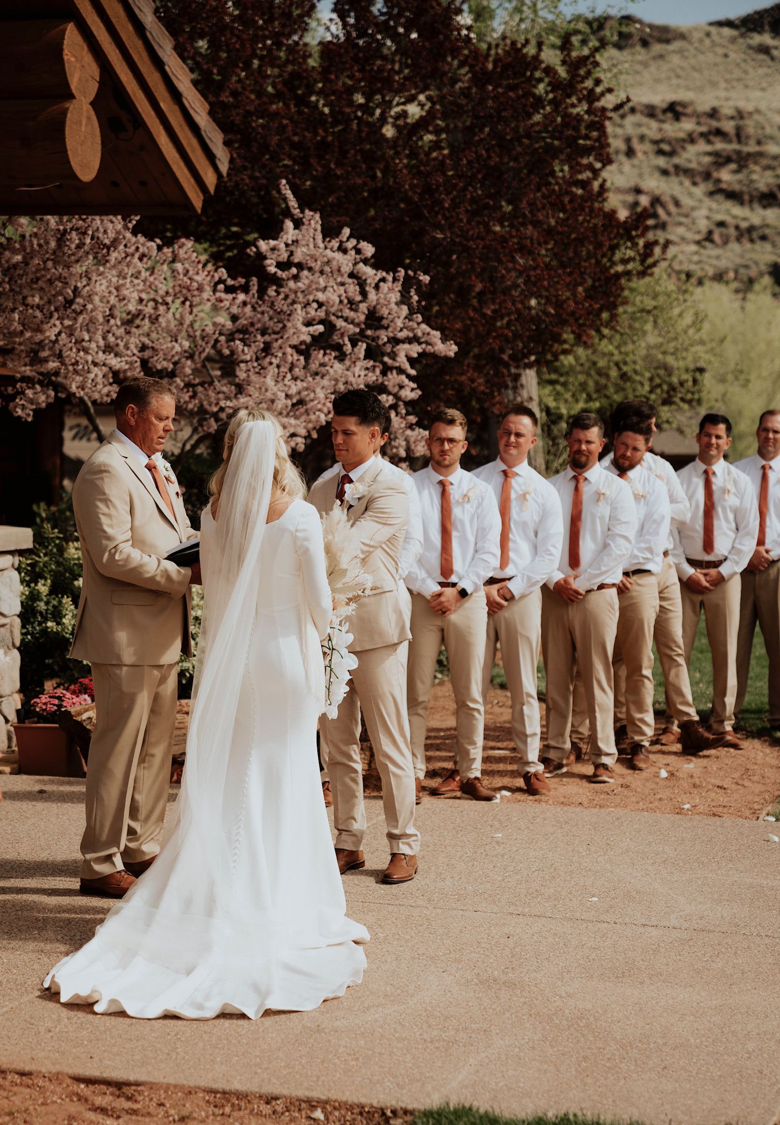 Wedding ceremony under a floral arch, bride in white facing groom and groomsmen in tan suits outdoors