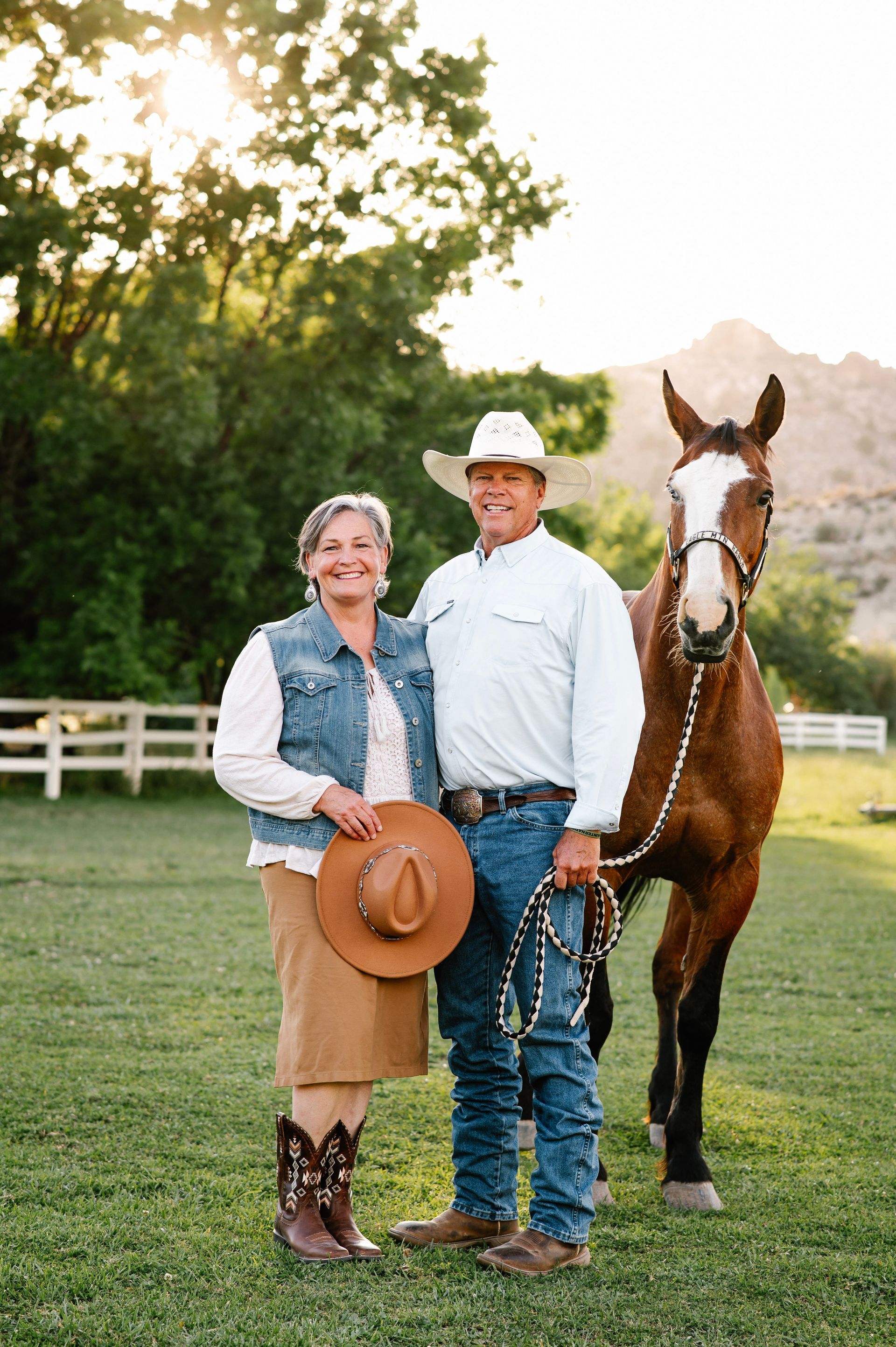 Two people standing with a brown horse in a grassy field, one holding a hat and the horse wearing a bridle.