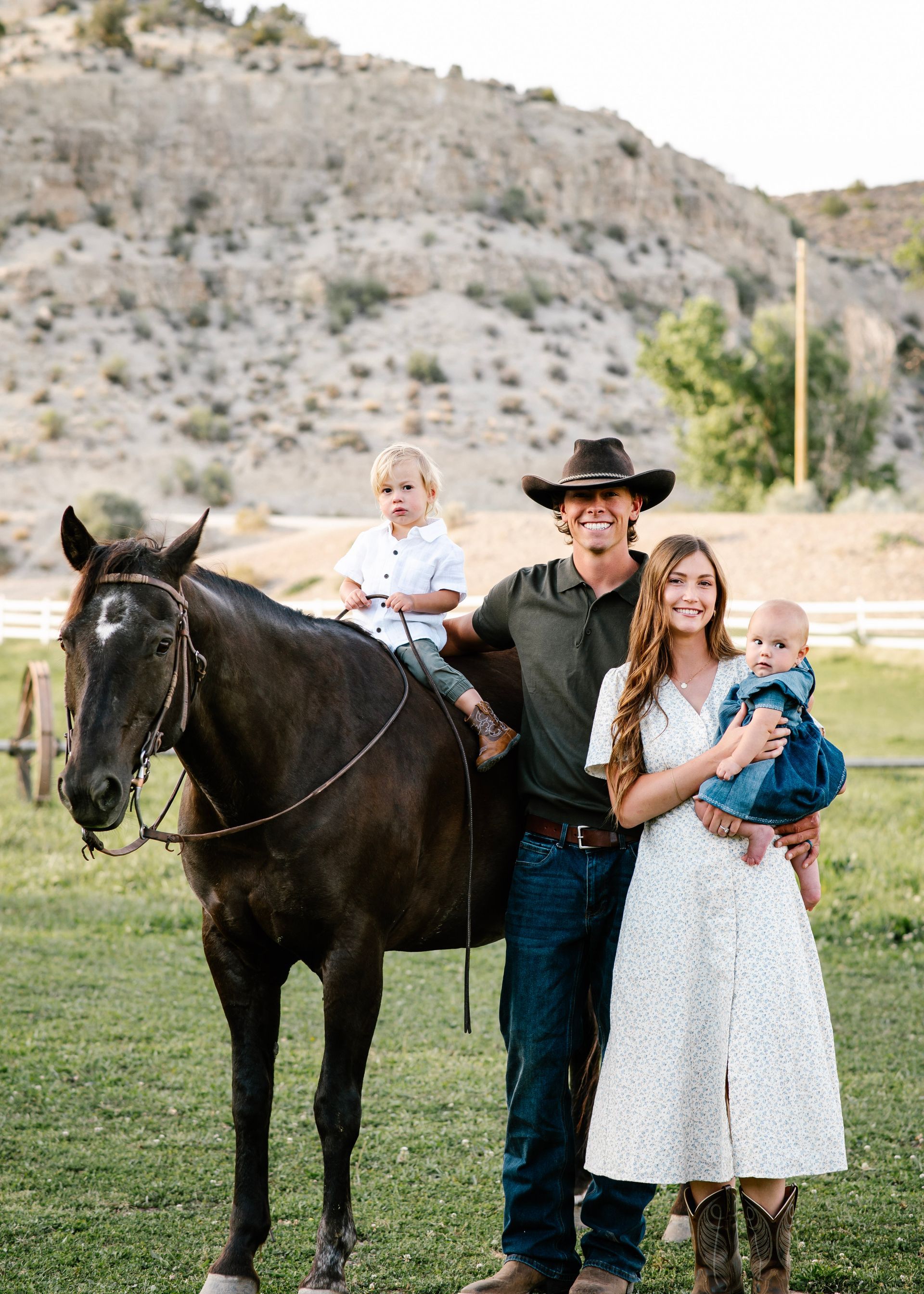 Family posing with a black horse in a grassy field, with rocky hills in the background.