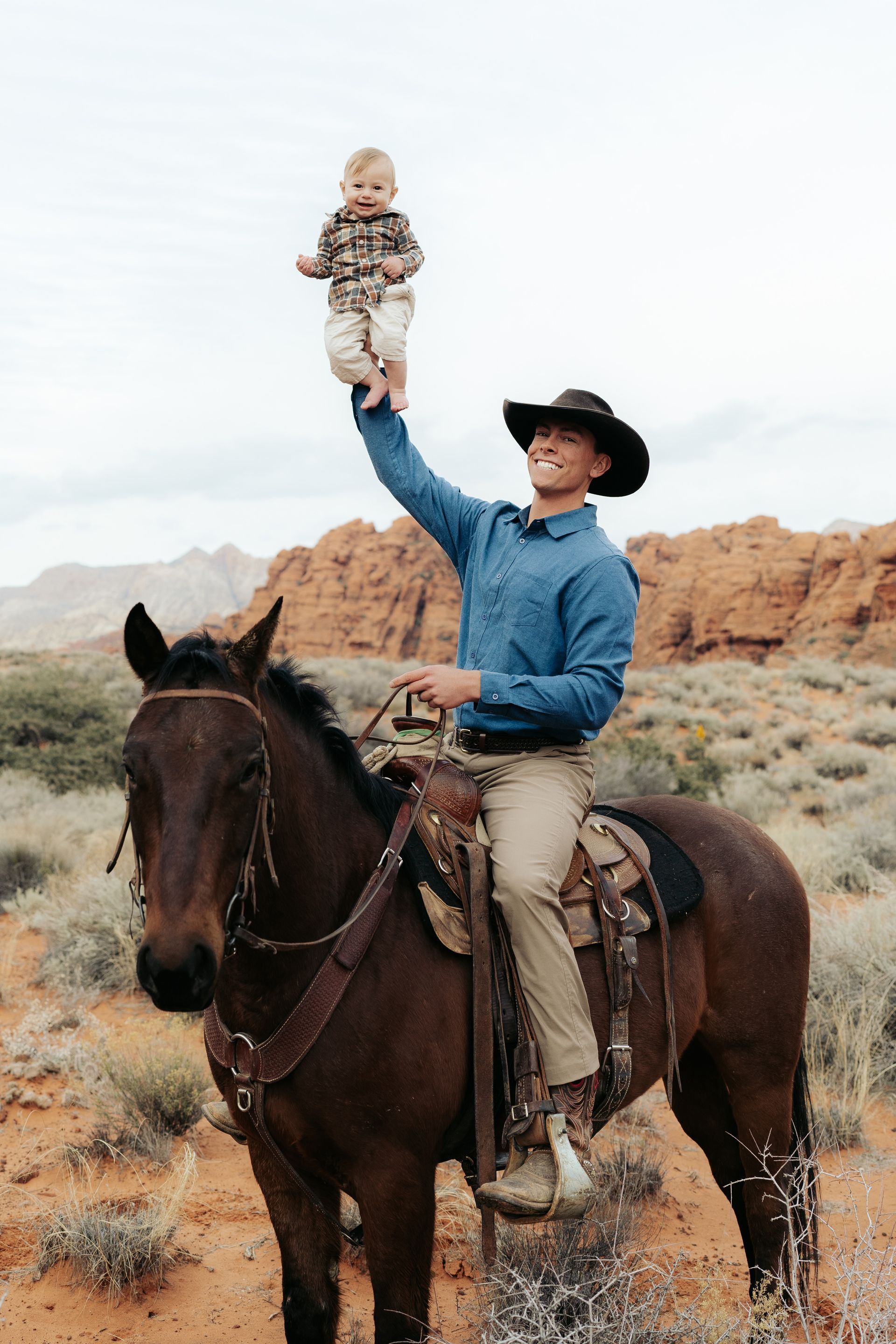 Cowboy on horseback lifting a baby in a desert landscape with rocky cliffs.