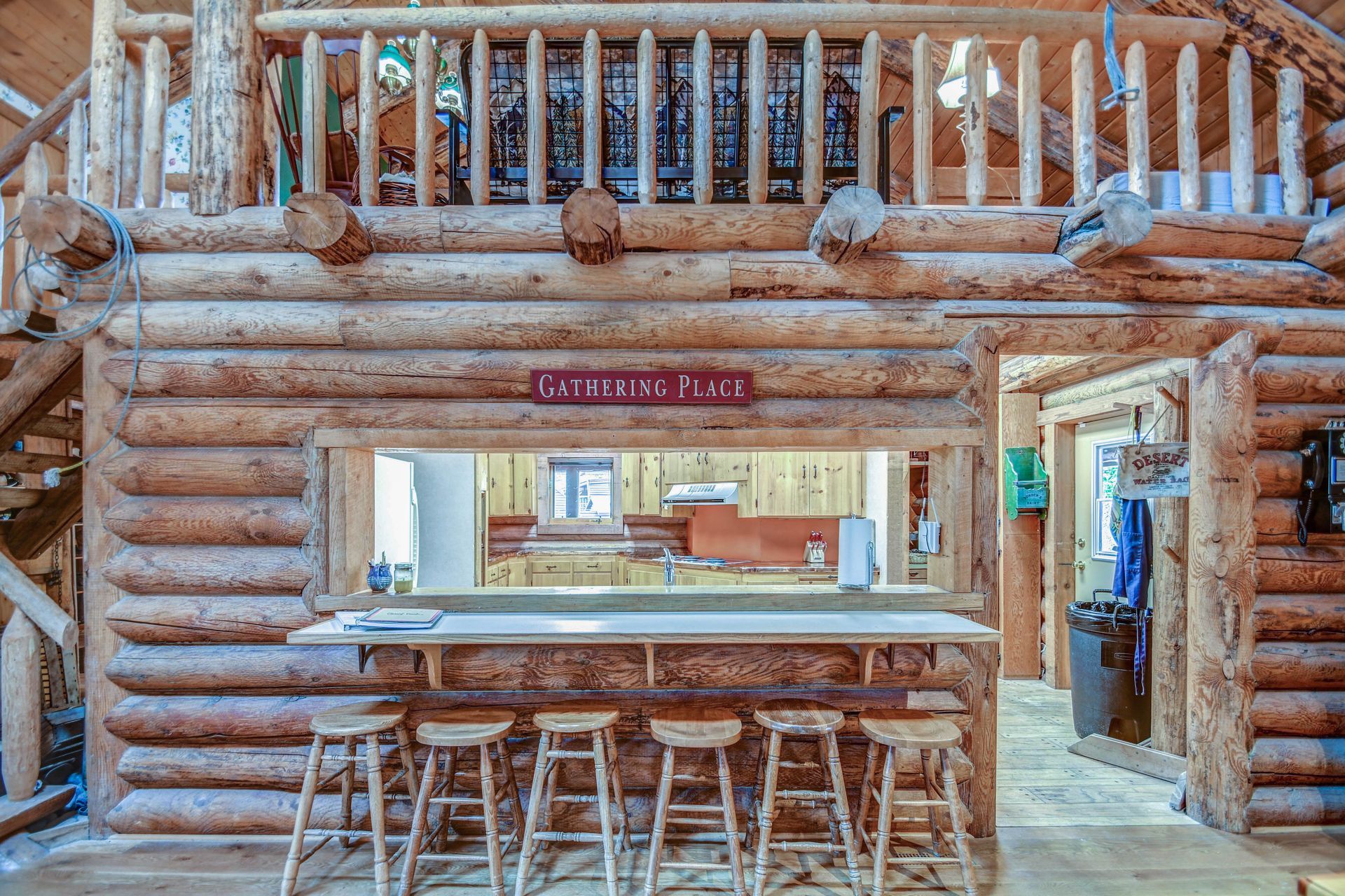 Rustic log-cabin bar with wooden stools, a counter window, and a balcony above