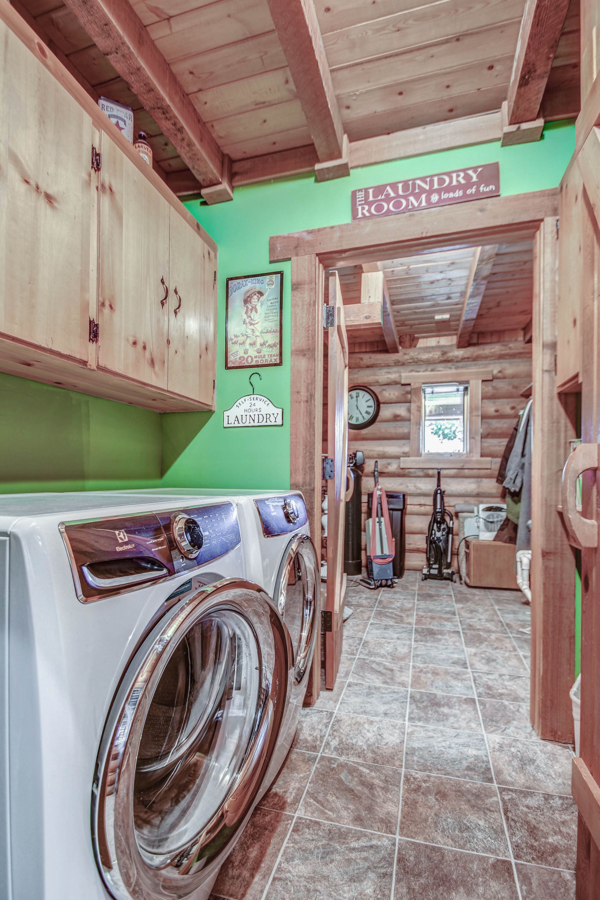 Narrow laundry room with washer, wood cabinets, green walls, and a tiled floor leading to another room