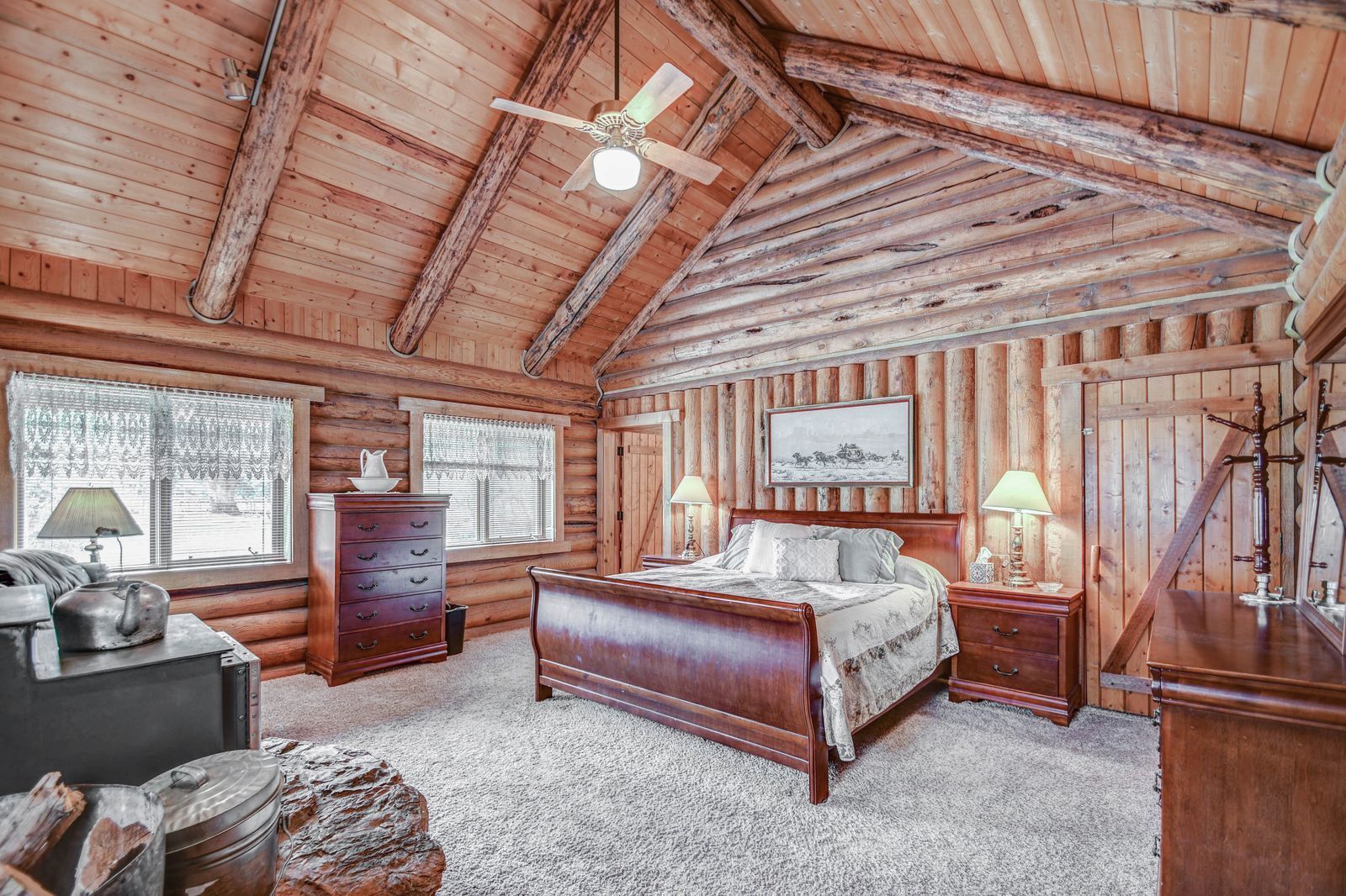 Rustic log cabin bedroom with a wooden bed, dresser, lamps, and vaulted ceiling with ceiling fan
