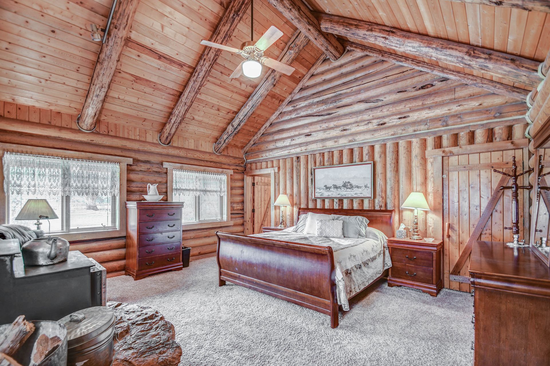 Rustic log cabin bedroom with wood walls, vaulted ceiling, wooden bed, dresser, and bedside lamp.