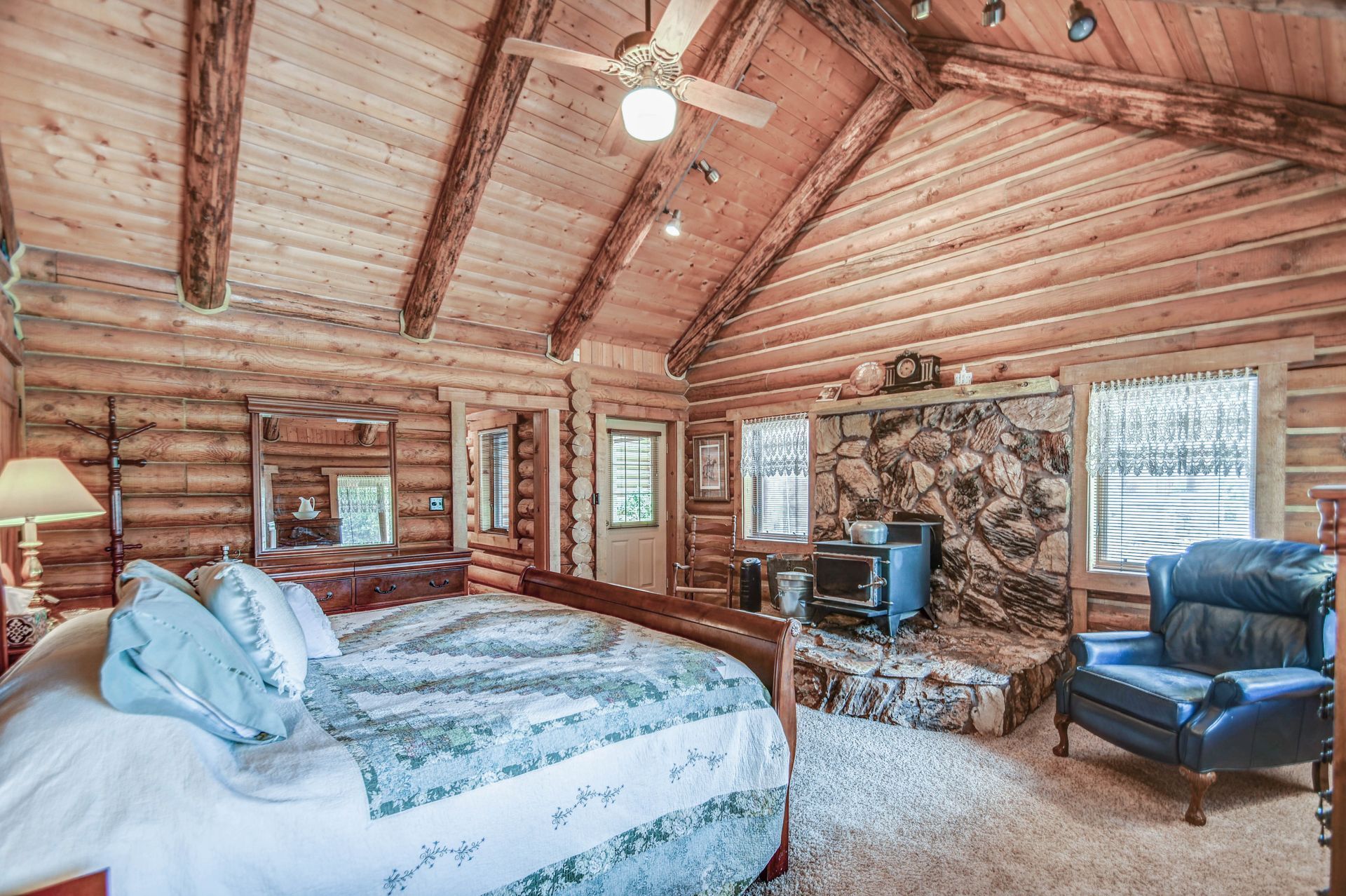 Rustic log cabin bedroom with a bed, armchairs, stone fireplace, and large windows