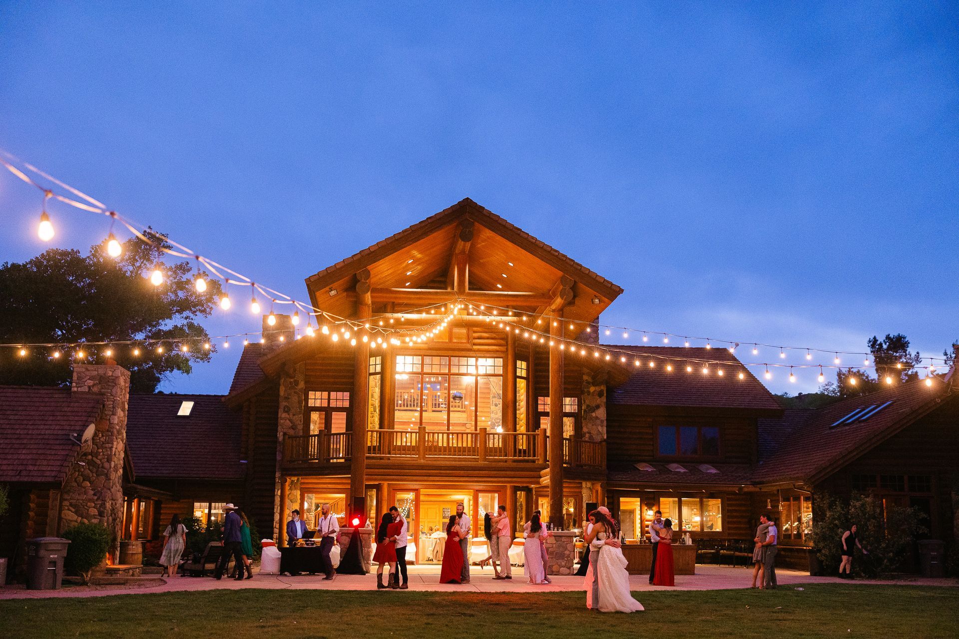 Lit wooden lodge at dusk with string lights, people gathered outside, and a couple standing in front.