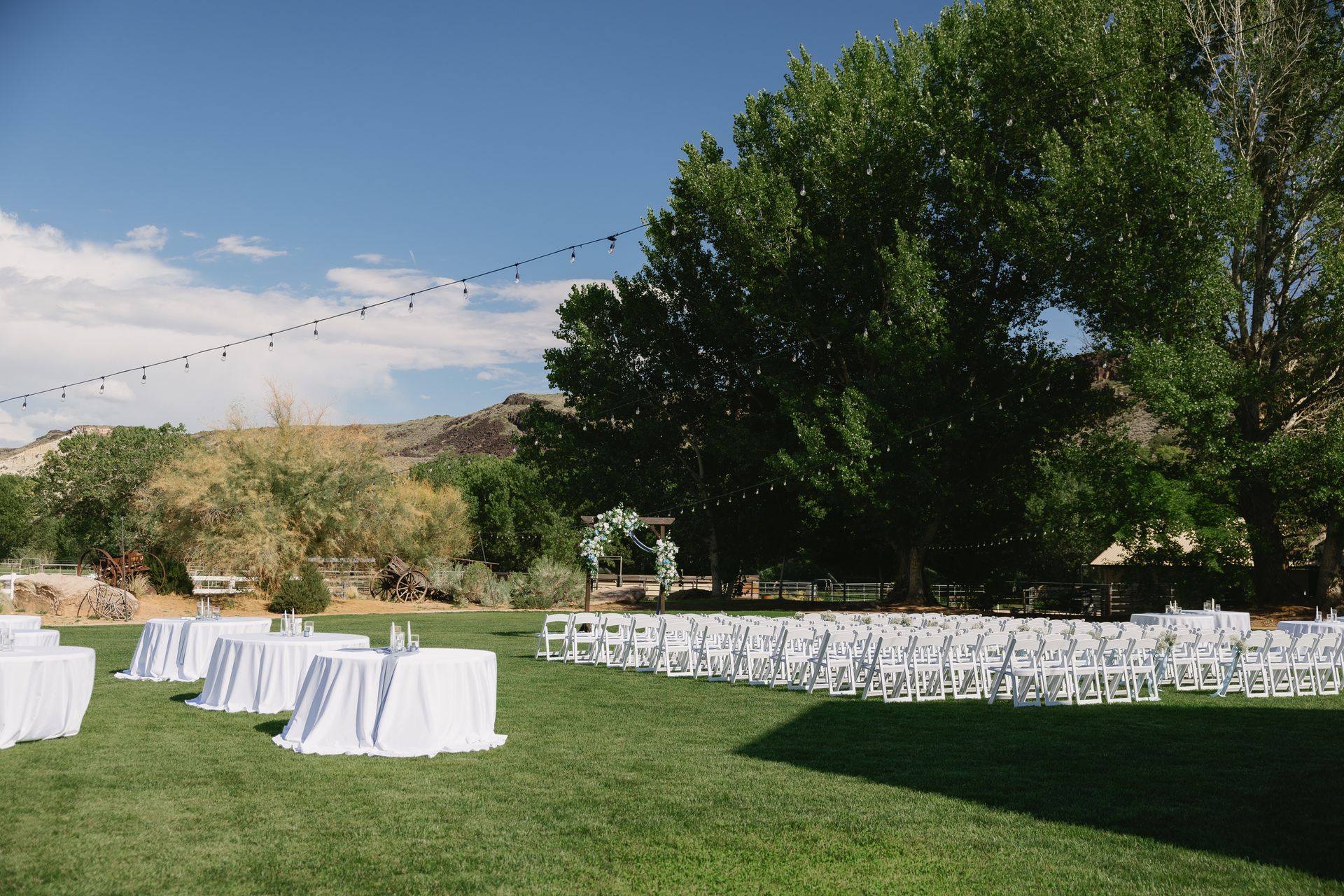 Outdoor wedding setup on a grassy lawn with white tables, chairs, string lights, and trees in the background