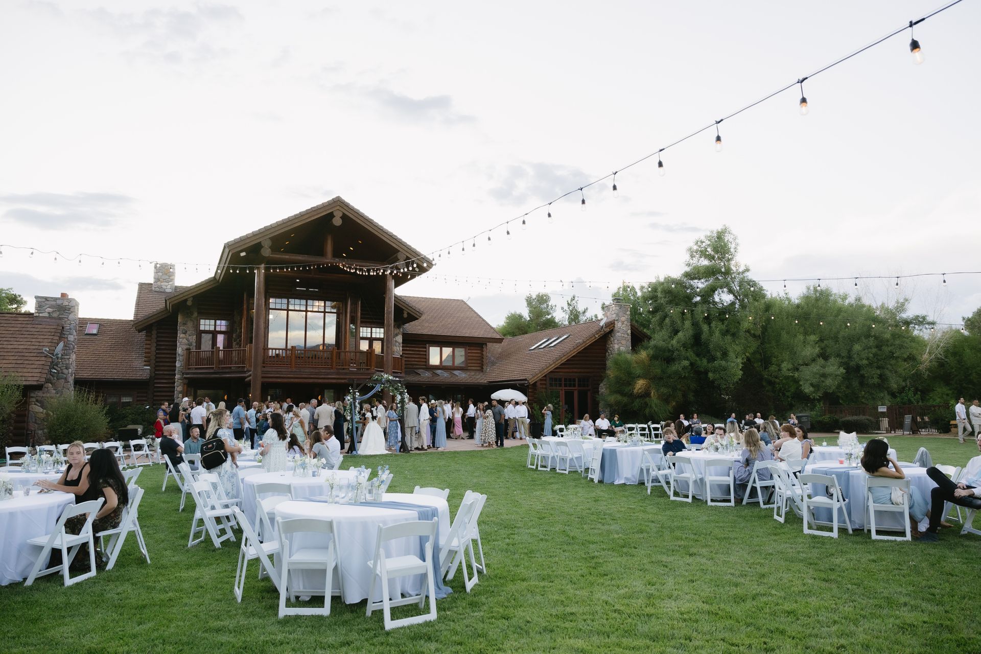 Outdoor wedding reception on a grassy lawn with white tables, guests, and a rustic barn backdrop
