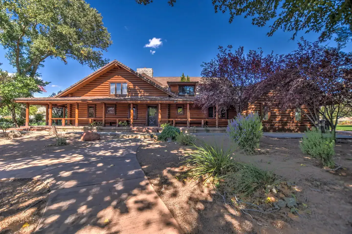 Rustic log cabin home with a front porch, surrounded by trees and desert landscaping under a blue sky