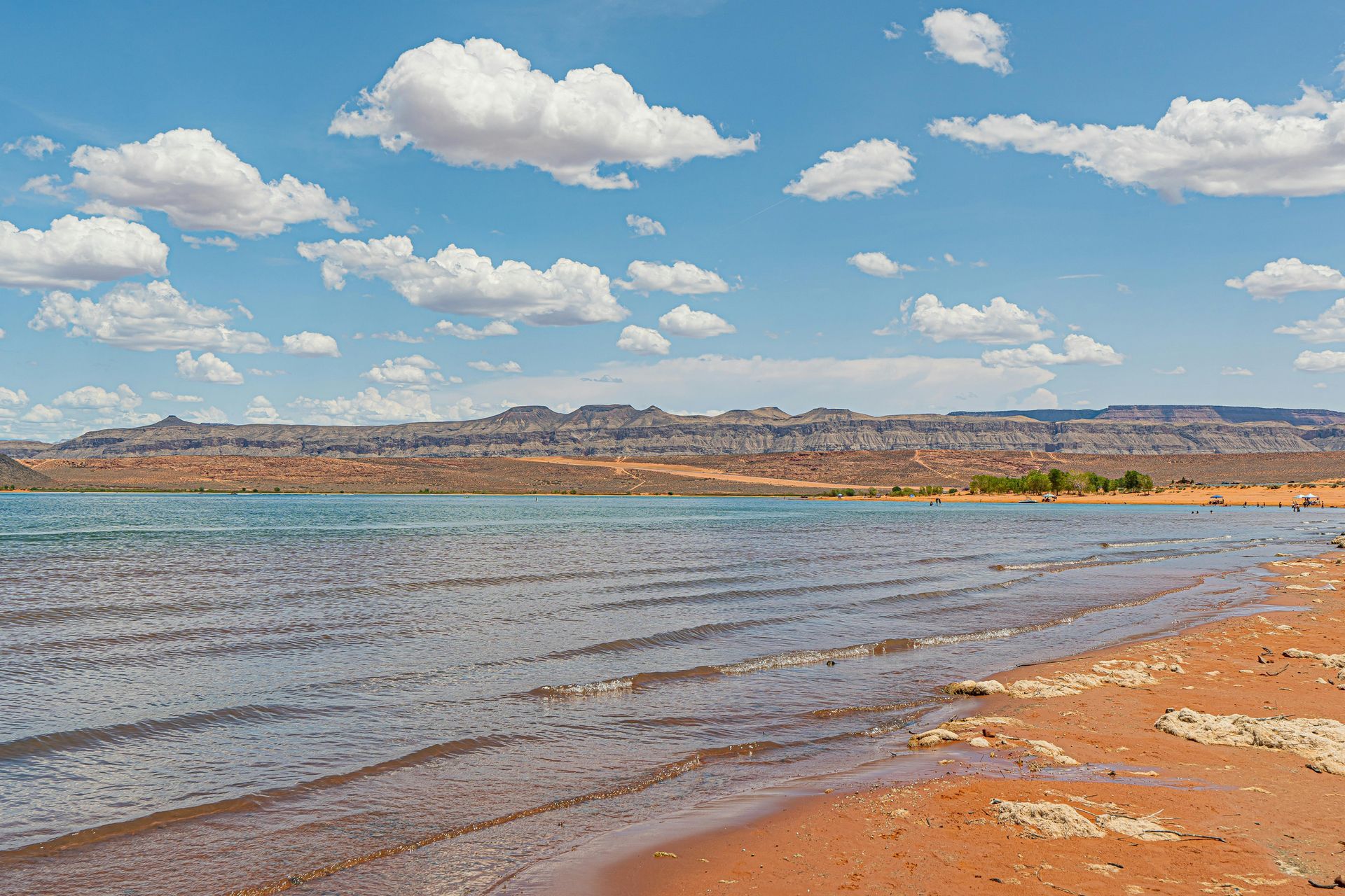 Sandy lakeshore with calm blue water, distant hills, and a bright sky with scattered clouds