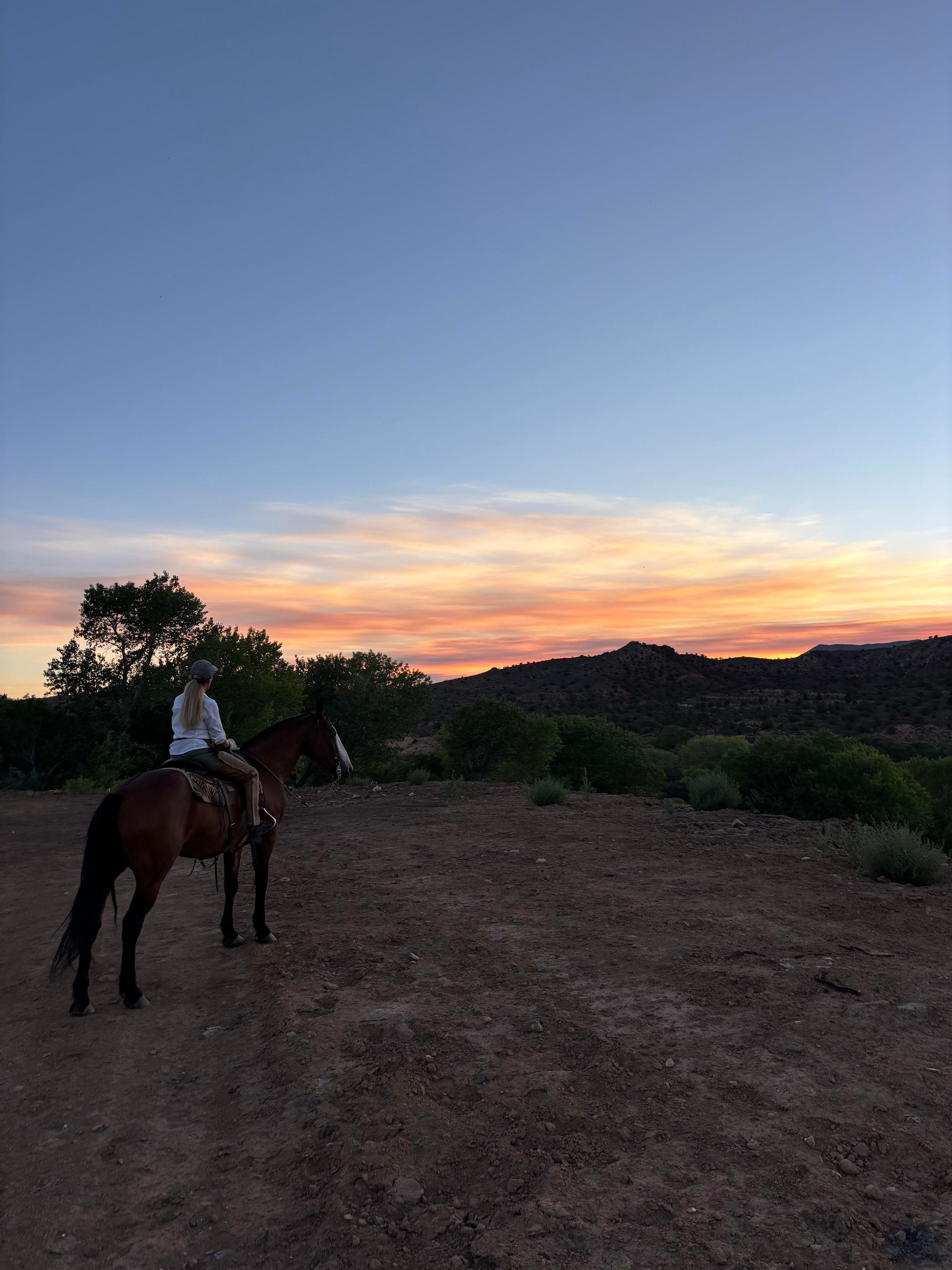 Person riding a horse on a dirt path at sunset with a colorful sky