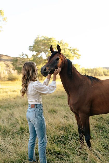 Woman in jeans gently touching a brown horse in a grassy field at sunset