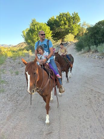 Two people riding horses on a dusty trail, with green trees and hills in the background.