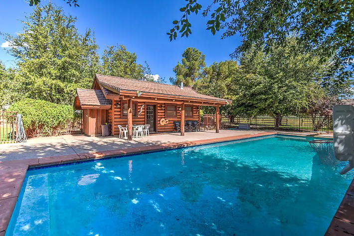 Backyard swimming pool beside a wooden cabin, surrounded by trees on a sunny day
