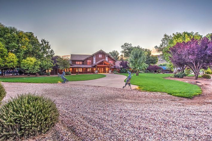 House with lit windows beside a gravel driveway and green lawn, framed by trees at dusk
