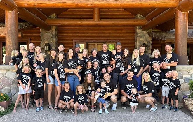 Large group posing in front of a log cabin, many wearing matching black shirts.