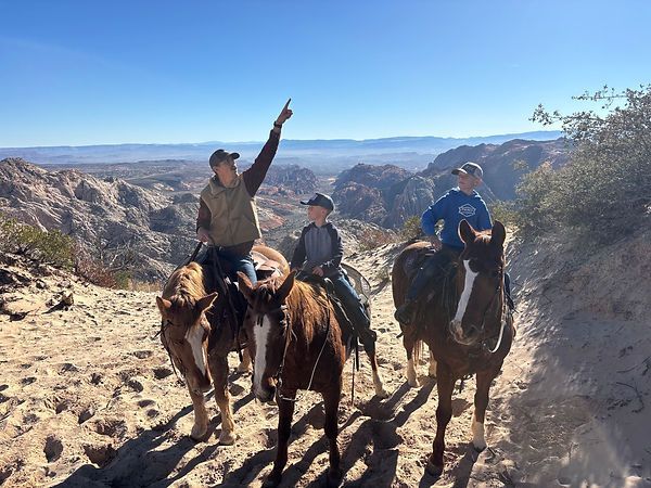Three horseback riders on a canyon trail, one pointing toward a bright desert valley.
