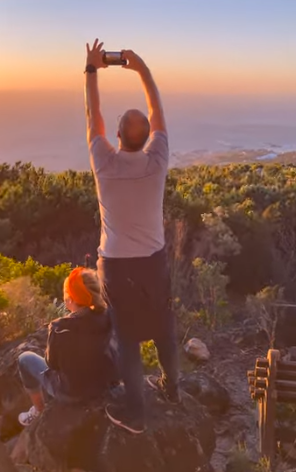 Two hikers on a hilltop at sunset, one holding a phone overhead above the landscape.